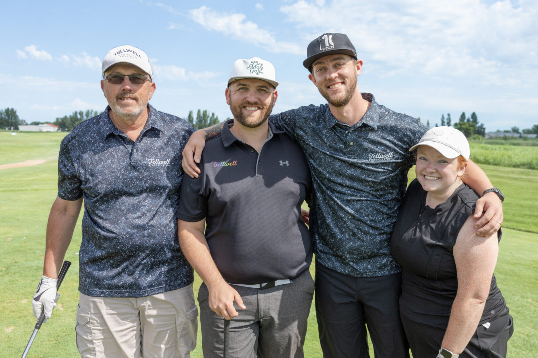 Three men and one woman stand on a gold course and pose for a photo. They are all wearing golf polos and hats. The sky is blue and slightly cloudy.