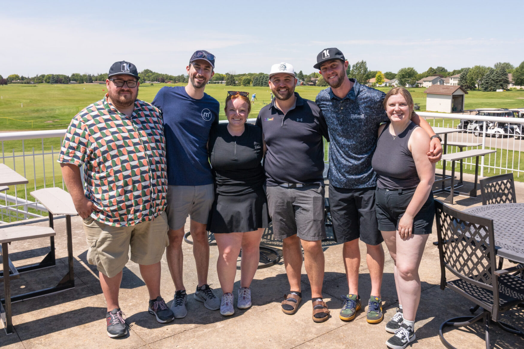 A group of six adults stands closely together, smiling for a photo on a sunny patio overlooking a golf course. The group includes four men and two women, dressed in casual summer clothing, some wearing golf attire and caps. Behind them are metal patio tables and chairs, as well as a view of the lush green course, golf carts, and a few buildings in the distance. The mood is cheerful and relaxed.