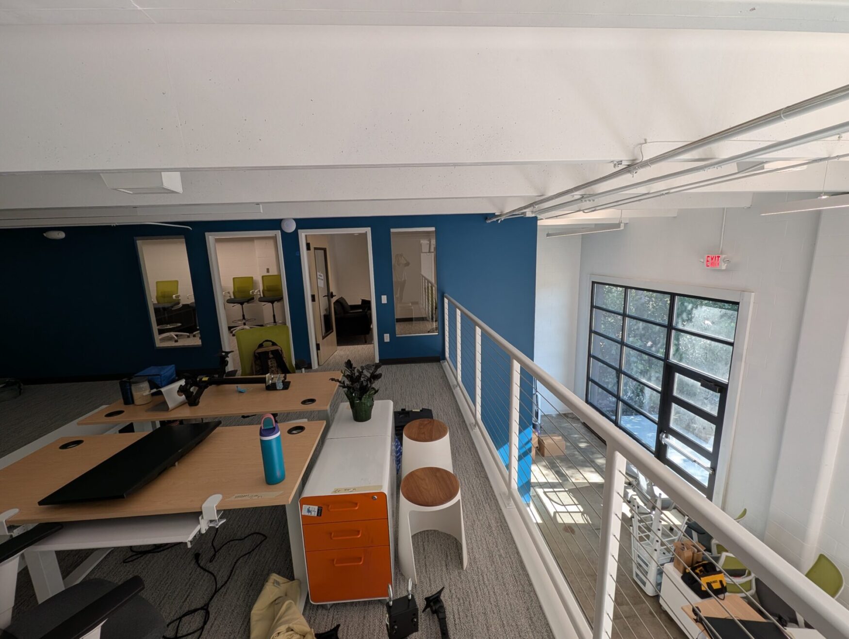 A modern, open-concept office space with a mezzanine level is shown. The foreground includes shared desks with a few scattered items like a monitor, water bottle, plant, and orange rolling drawer. To the right is a railing overlooking the lower floor, which has large windows and a glass garage-style door letting in natural light. In the background, there are three office rooms with glass doors and windows, one of which contains bright green chairs around a table. The walls are painted blue and white, and the flooring is a light-colored carpet. The space appears to be in the process of being set up or reorganized.