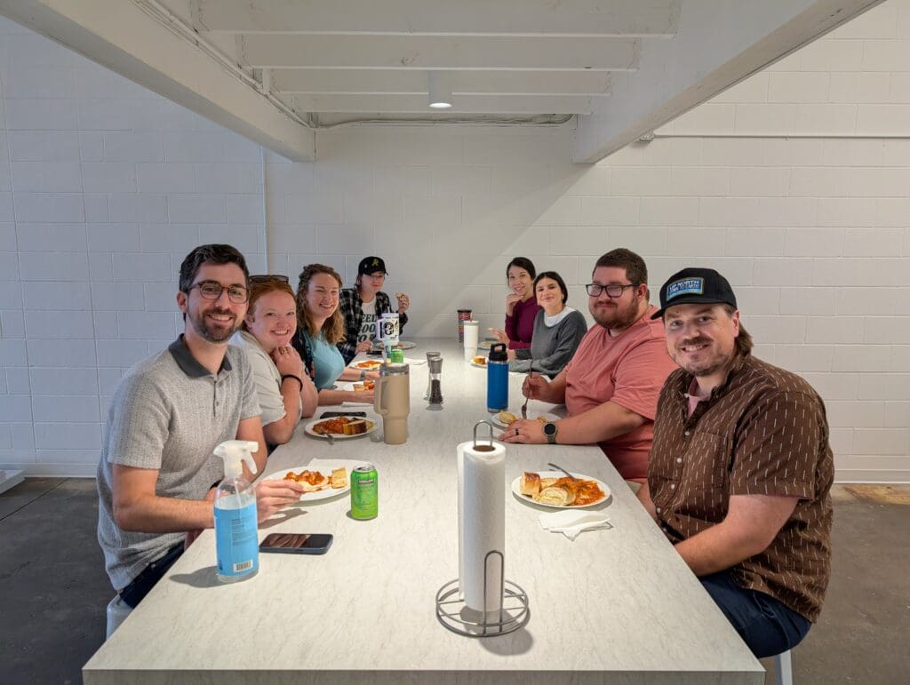 A group of 8 men and women sit at a large table sharing a meal. There are plates with pasta and garlic bread, an array of drink cans, and cleaning supplies on the table as well.