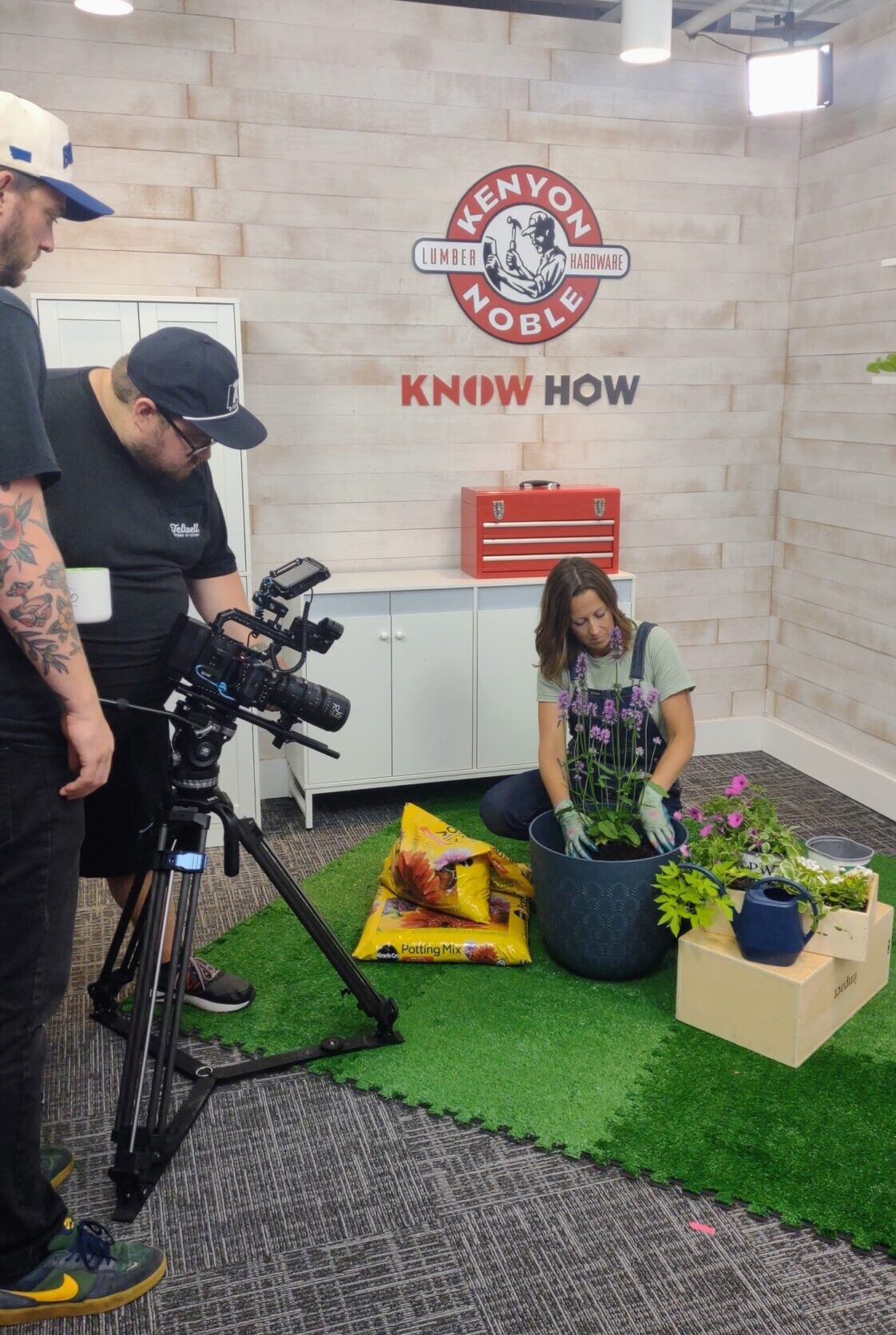 A video production setup captures a woman planting flowers in a studio decorated with a hardware store theme. She kneels on artificial grass, surrounded by potted plants, potting soil bags, and gardening tools. Two camera operators film the scene under studio lights, and a "Kenyon Noble" lumber and hardware logo is displayed on the wall behind her.