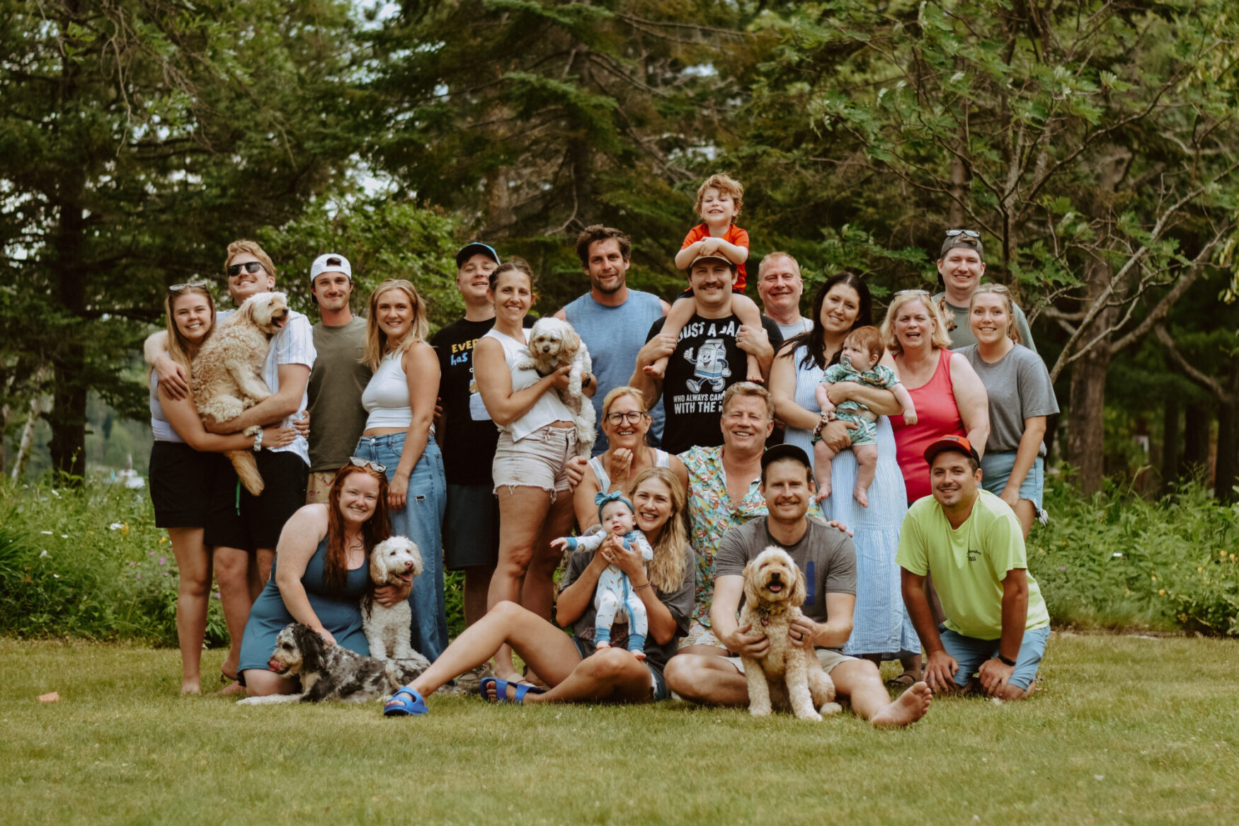 A large multigenerational family poses outdoors on a grassy lawn surrounded by trees. The group includes around 25 people of various ages—from babies to older adults—along with several dogs. Most people are smiling and dressed in casual summer clothing. Some are sitting on the ground, some are kneeling, and others are standing in rows. It's a relaxed and cheerful family gathering, likely during a summer reunion or vacation.