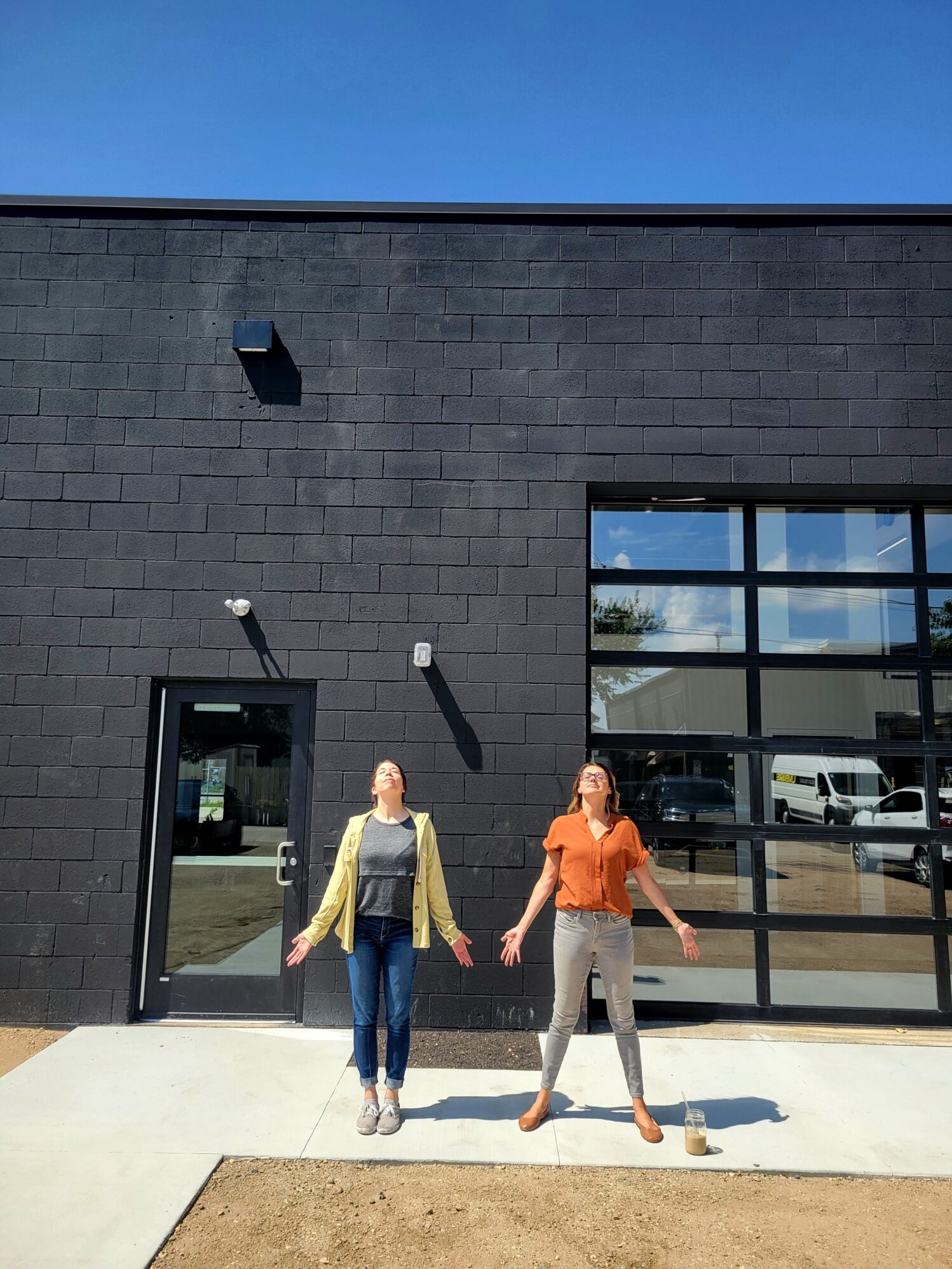 Two women stand with their faces angled towards the sun and their arms out next to them. They are enjoying the sunshine in front of a black building with a garage door.