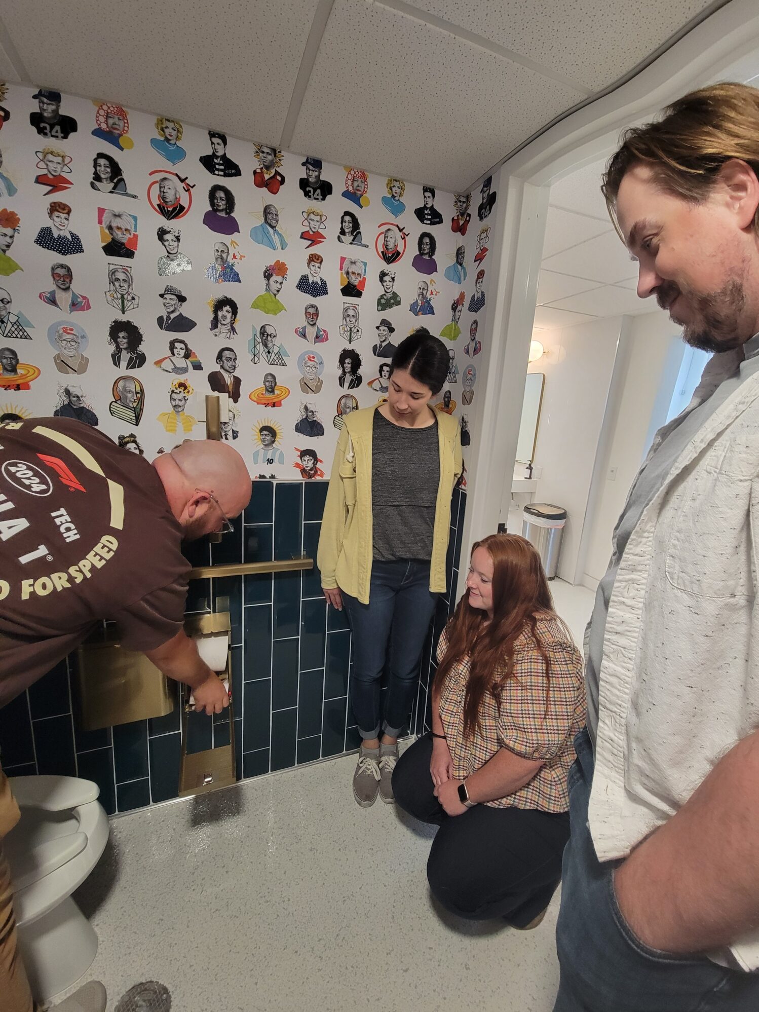 Four people gather in a bathroom with a colorful celebrity portrait wallpaper. One person kneels near the toilet paper holder, inspecting or demonstrating it, while the others watch and smile.