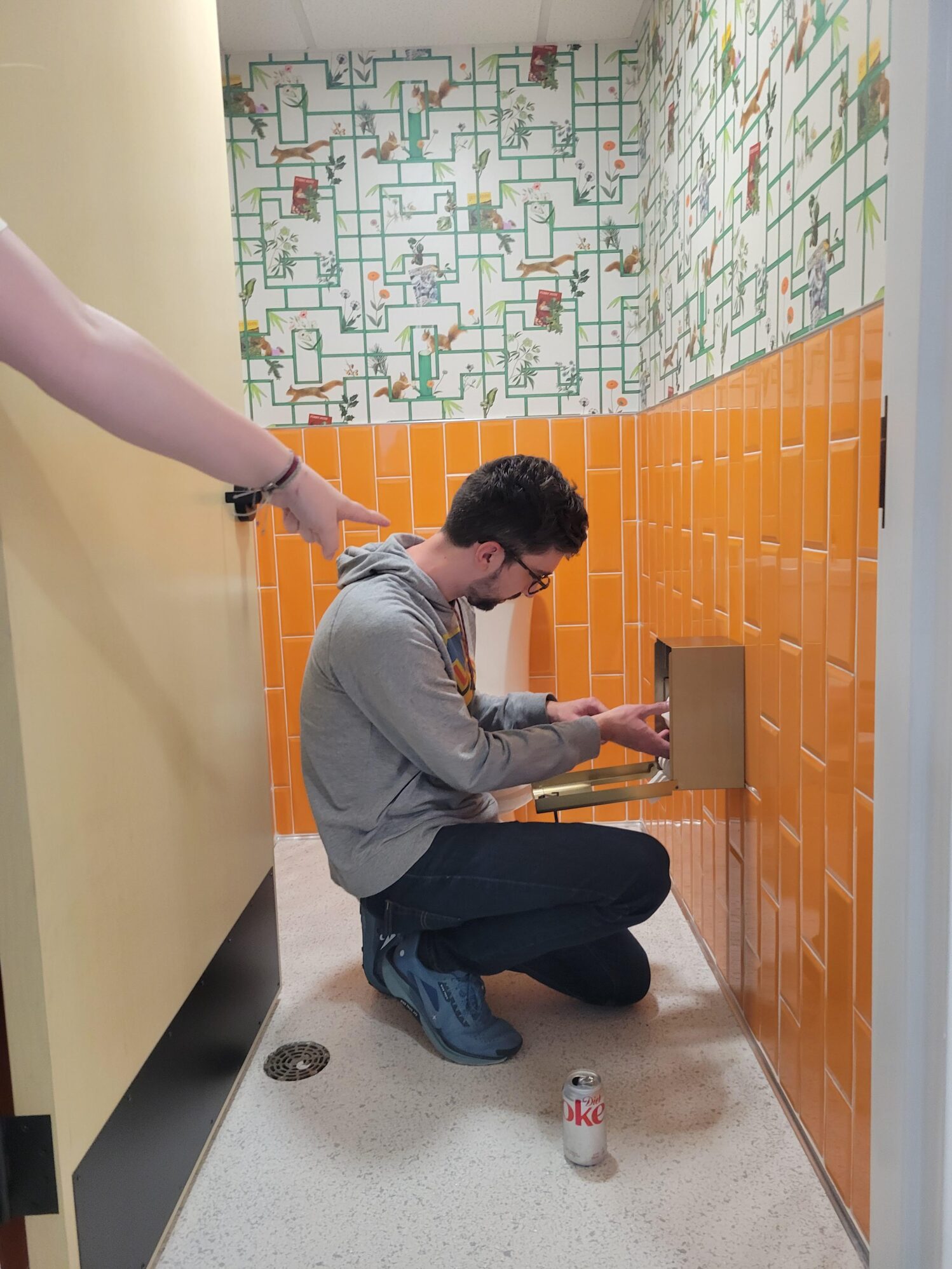 A person kneeling in a brightly tiled bathroom, adjusting a gold toilet paper holder. A can of Diet Coke sits on the floor, and another person’s hand points toward the scene.