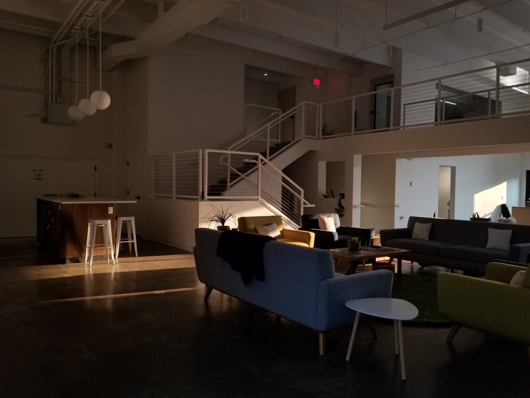 A modern loft-style office lounge area during golden hour, with warm sunlight streaming in from the right. The space features a mix of mid-century modern furniture, including gray, blue, and green armchairs and sofas arranged around wooden coffee tables. A staircase with white railings leads to an upper-level walkway with office rooms. In the left corner, there's a kitchen island with two metal stools and spherical pendant lights hanging above. The concrete floor and industrial ceiling pipes add to the minimalist, contemporary feel of the space.