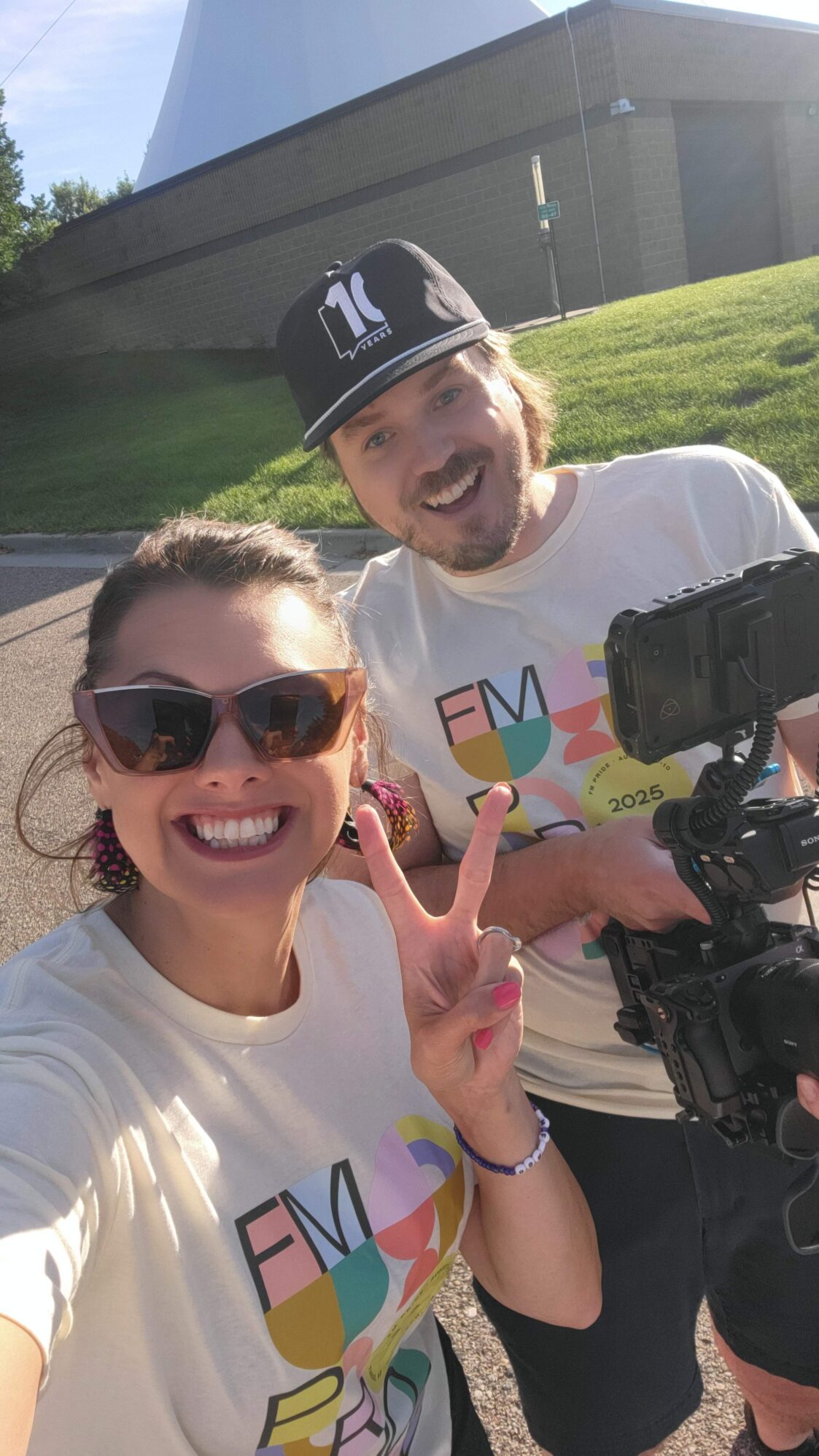 A woman in sunglasses makes a peace sign and smiles widely while taking a selfie with a man holding a professional camera. Both wear matching T-shirts with a colorful design, standing outside in the sunshine near a large white structure.