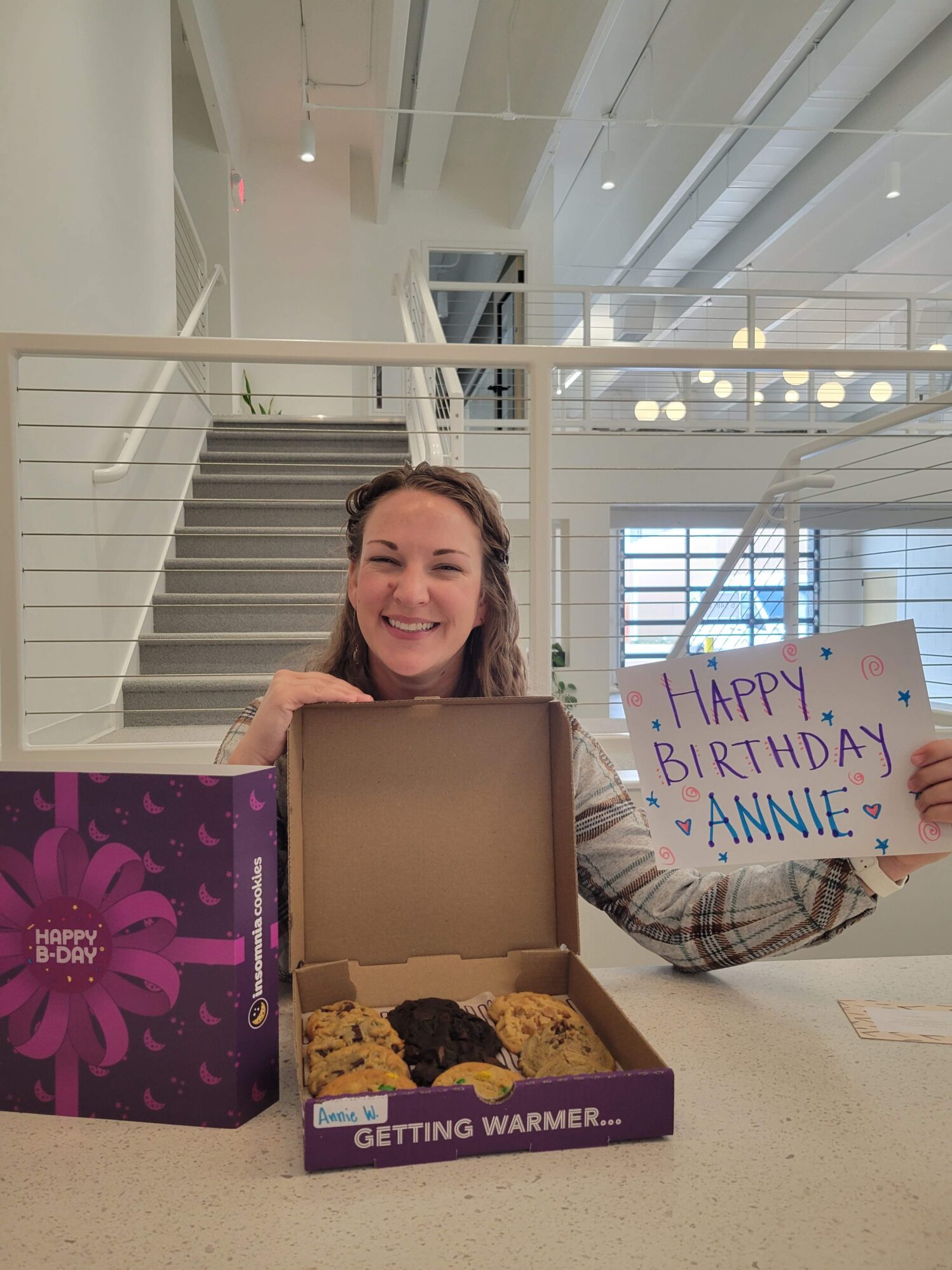 A woman holds a sign that reads “Happy Birthday Annie!” She is also holding open a cardboard box filled with an assortment of cookies. She is in an industrial office setting.