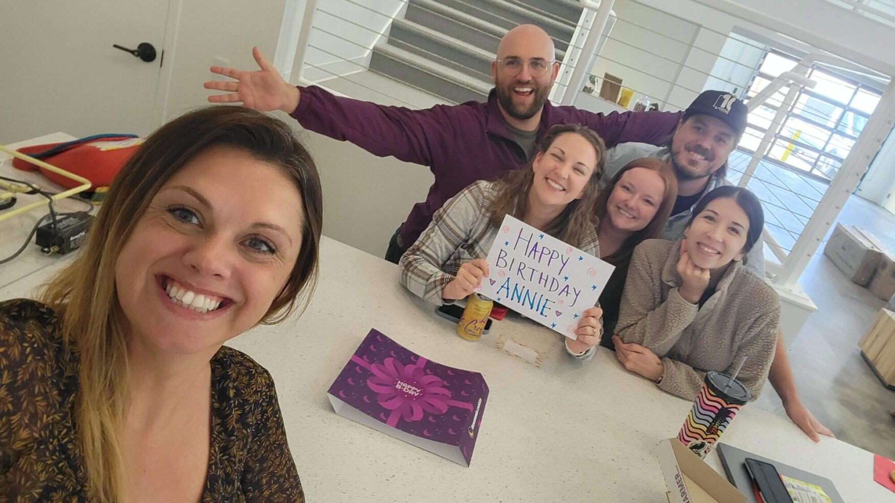 A group of six people stand in the kitchen of an office space, all smiling for the camera. One woman holds a sign that reads “Happy Birthday Annie!”