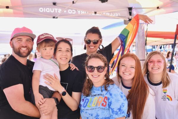 A group of six people, including a young boy, smile under a Tellwell Story Co. tent at a Pride event. Several are wearing rainbow-themed shirts, one person holds a rainbow flag, and everyone looks cheerful and celebratory.