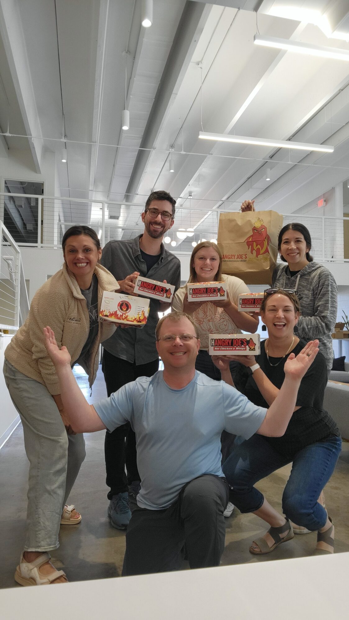 A group of six people pose for a photo in a bright office space. Five of them are holding boxes that read “Hangry Joe's Hot Chicken & Wings”. One man is kneeling in the front of the group with both arms up, showing off the boxes.