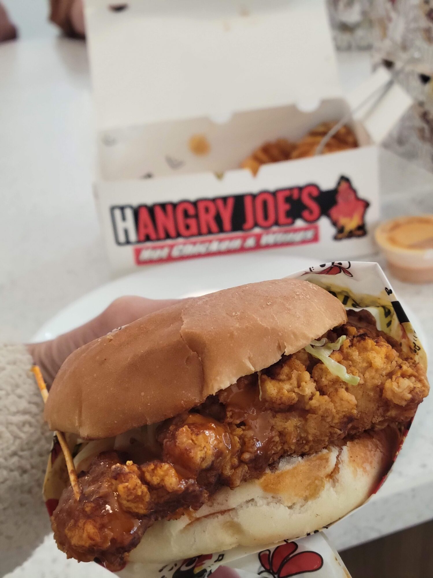 A fried chicken sandwich is shown, with the meal box in the background. The box reads “Hangry Joe’s Hot Chicken & Wings”.