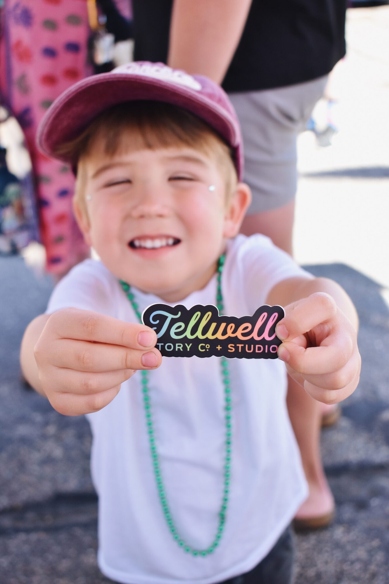 A young child wearing a maroon cap and green bead necklace holds a black and rainbow-colored “Tellwell Story Co + Studio” sticker toward the camera, smiling brightly in an outdoor setting.