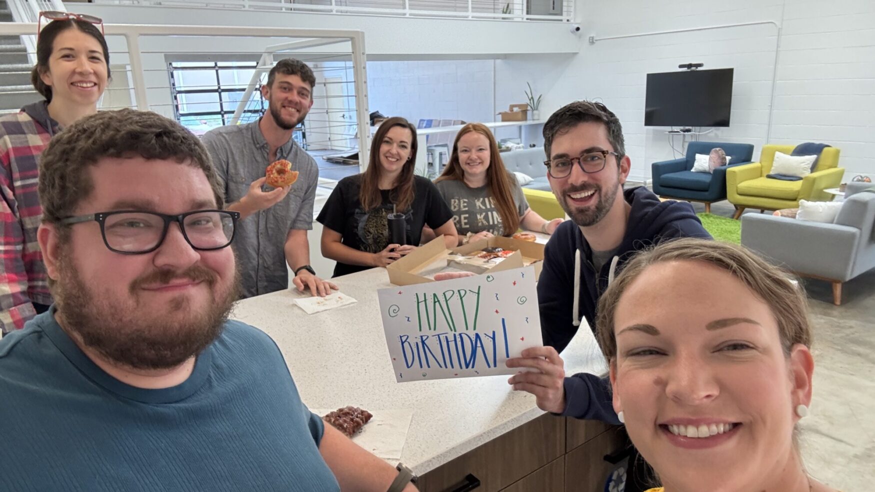 A group of seven people gathered around a kitchen island in a modern, open office space, smiling at the camera. One person holds a colorful handwritten sign that says 'Happy Birthday!' while others enjoy donuts. The atmosphere is cheerful and celebratory.