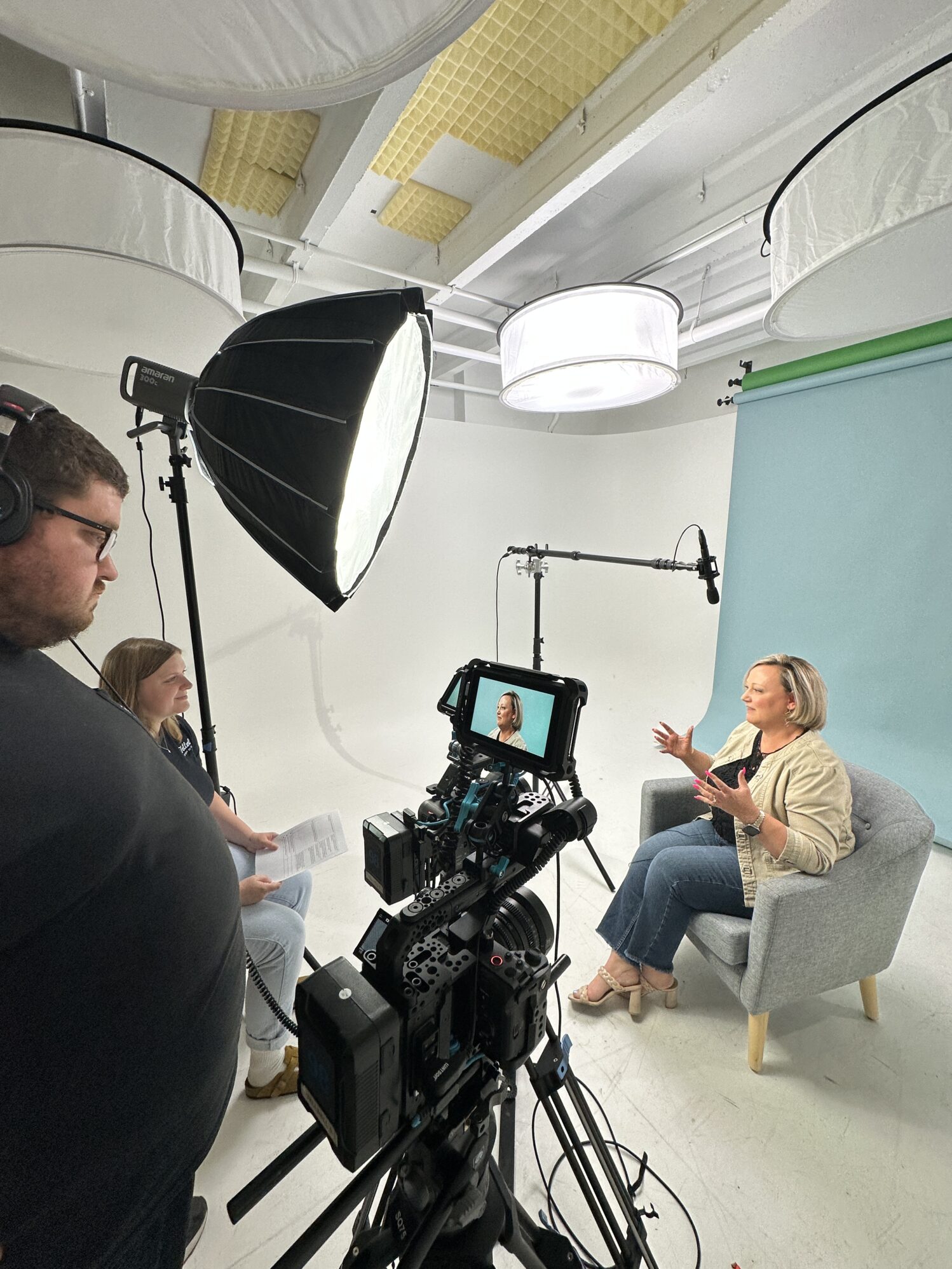 Behind-the-scenes view of a video interview setup in a studio. A woman sits in a gray armchair against a light blue backdrop, speaking with animated hand gestures. A camera on a tripod records her, and a monitor shows her framed shot. Two crew members sit nearby—one wearing headphones and one holding papers. Softbox and overhead lights illuminate the scene.