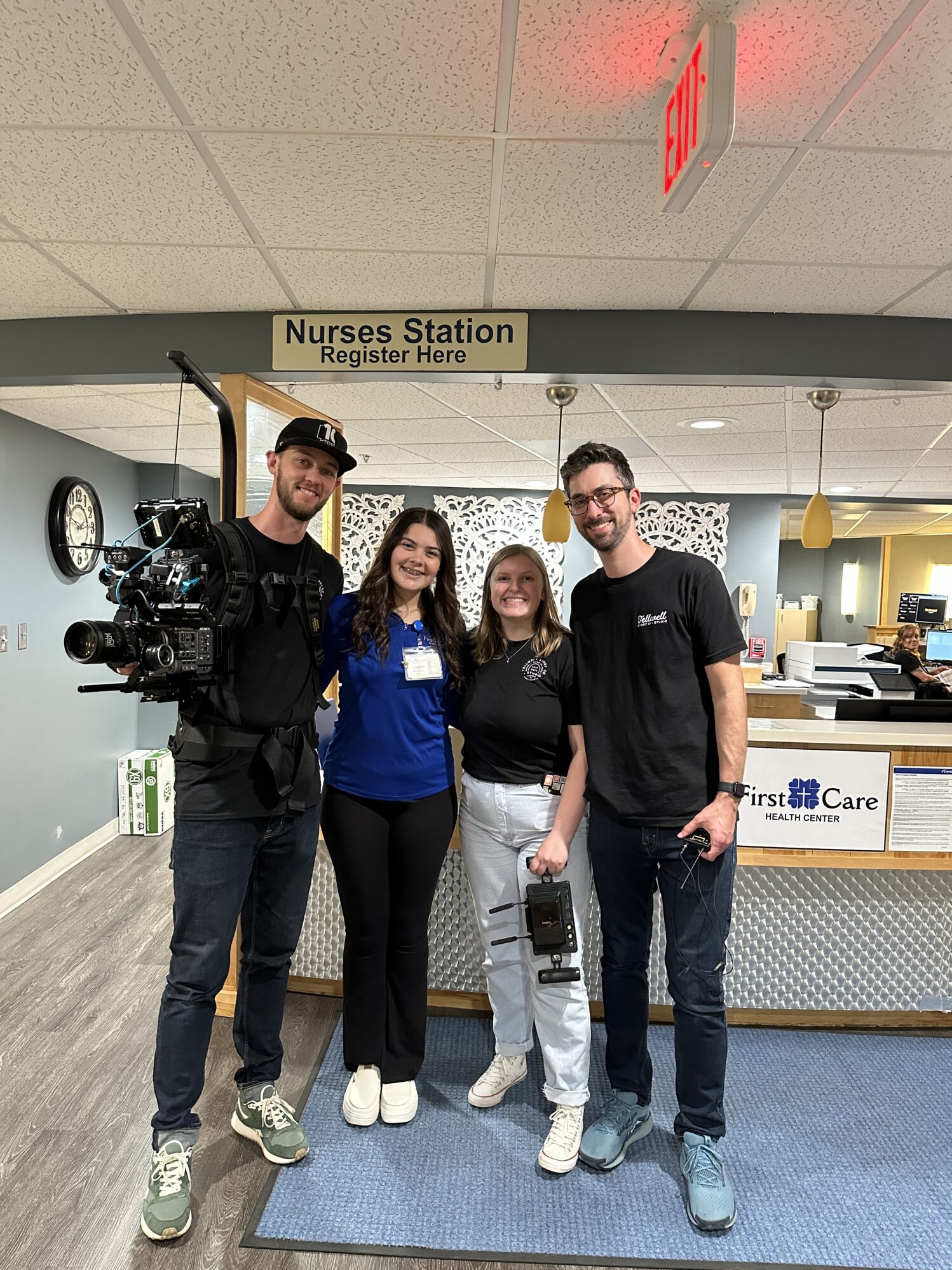Four people standing together and smiling inside a medical center by the nurses’ station. Two of them are holding professional video equipment, suggesting a filming crew, while the other two are staff members. A sign overhead reads “Nurses Station Register Here,” and part of the reception desk is visible in the background.
