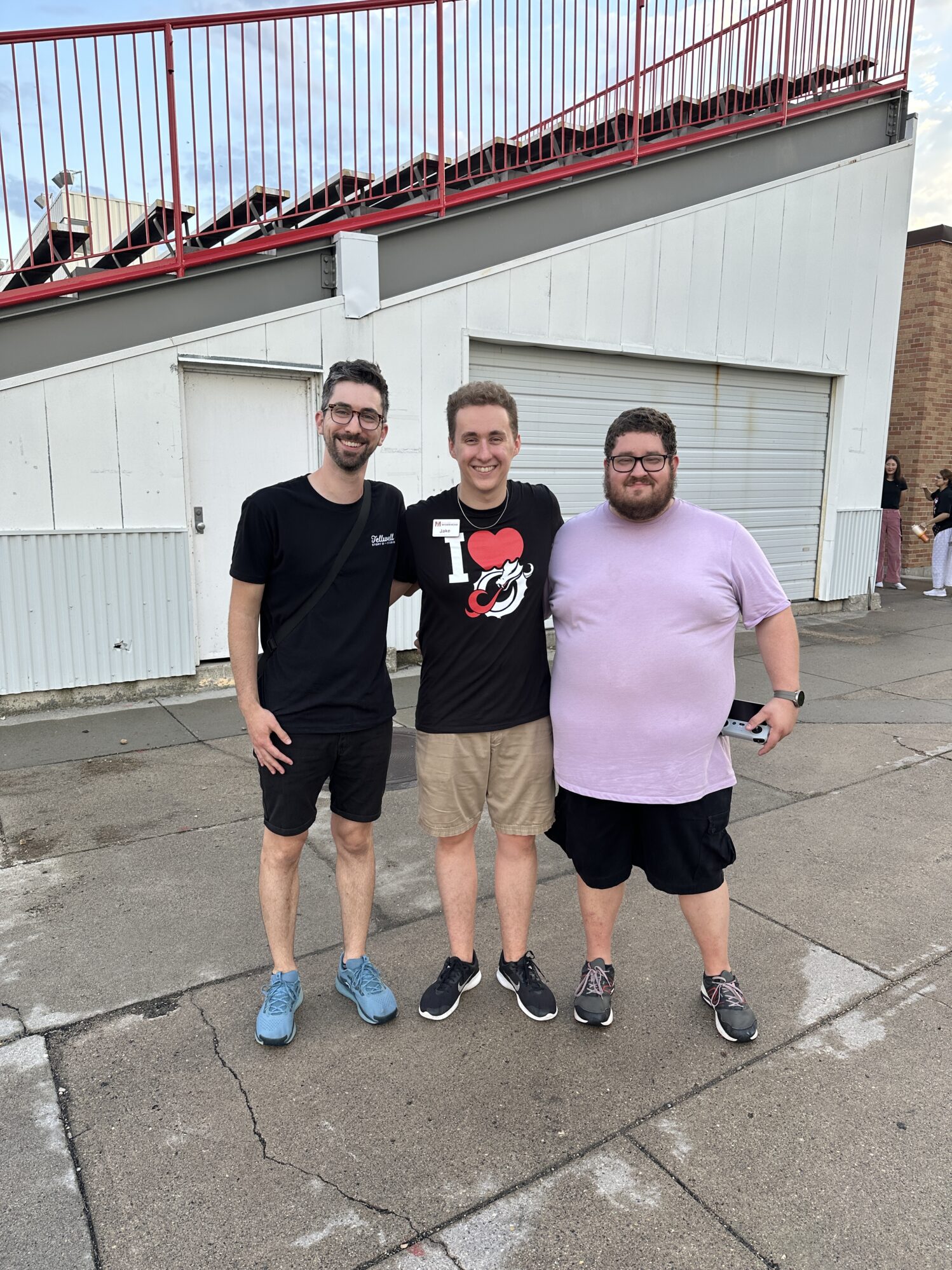 Three men pose for a photo. There are stadium bleachers in the background, suggesting they are at a football field.