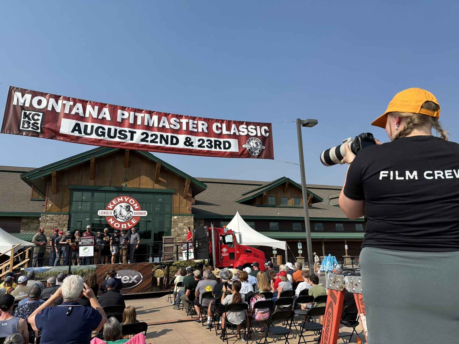A woman with a yellow hat and braids holds a camera, taking pictures at an outdoor event. There is a banner that reads “Montana Pitmaster Classic, August 22nd & 23rd.” There is a crowd sitting, watching an award ceremony. This is taking place in front of a large building with a sign that reads “Kenyon Noble Lumber & Hardware”.