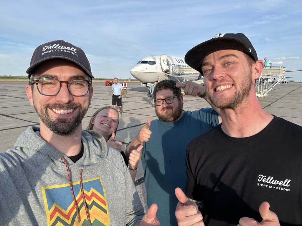 Three men and a woman stand on the tarmac at an airport with a plane in the background. They are all smiling for the camera and giving thumbs up.