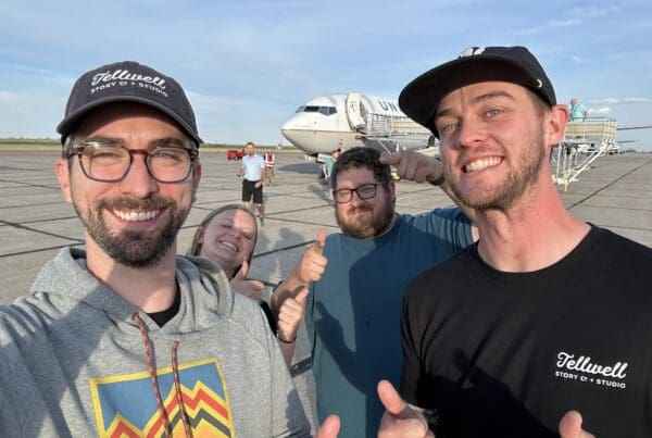 Three men and a woman stand on the tarmac at an airport with a plane in the background. They are all smiling for the camera and giving thumbs up.