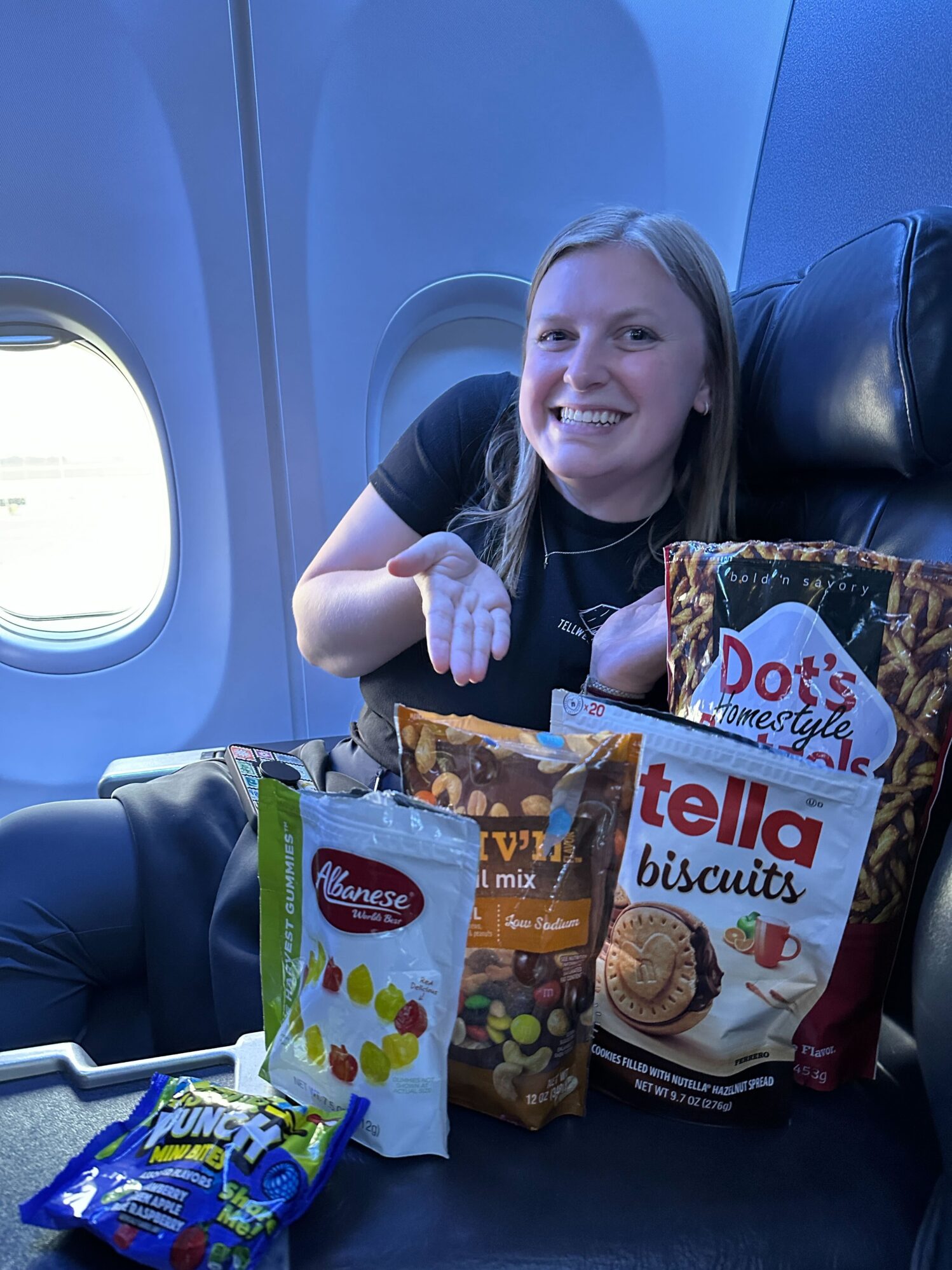 A woman sits in a plane seat with dim lighting. She is smiling and posing her hands to showcase several bags of snacks. There is a mix of gummy candy, cookies, and pretzels.