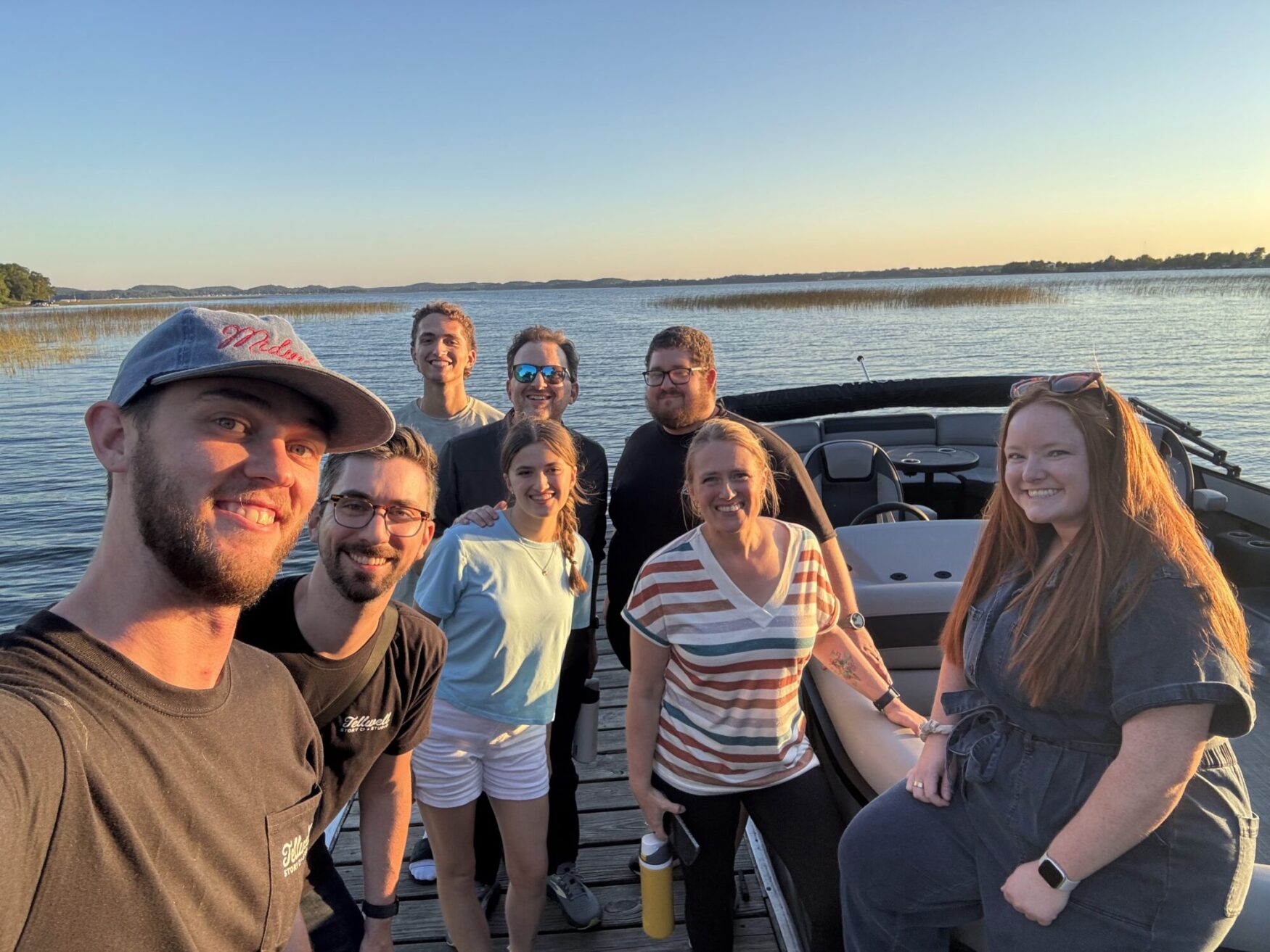 A group of people on a boat in the lake. The sun is setting, which is shining golden light onto their faces. They are all smiling for the photo and the atmosphere is happy.