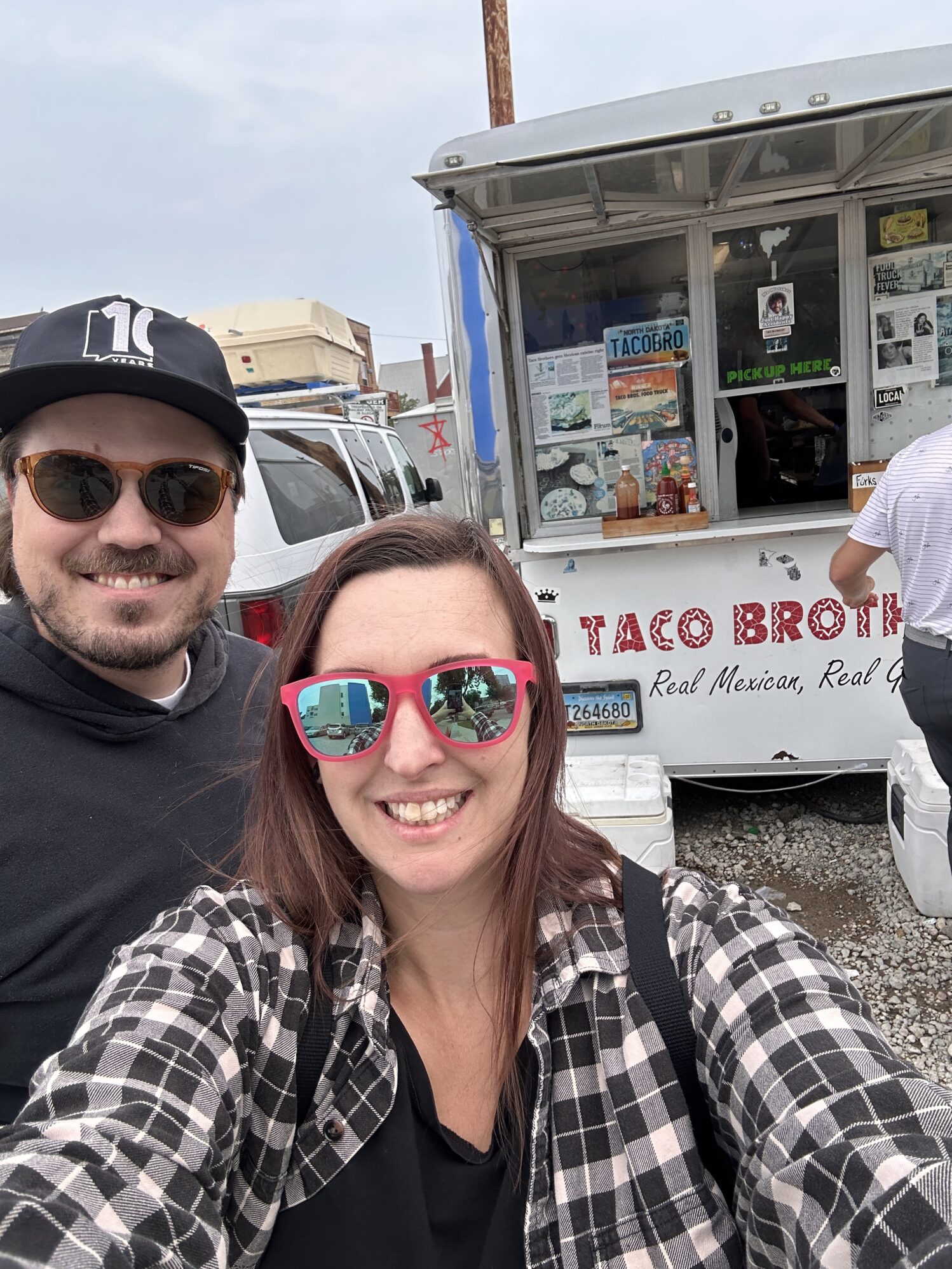 A man and woman pose for a selfie outside a food truck names "Taco Brothers". They are both smiling and wearing sunglasses.