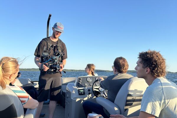 A group of people on a boat under a clear blue sky. One person is steering while others sit and talk. A cameraman with gear strapped to his chest is filming on deck. A yellow water bottle sits on a small table in the foreground.