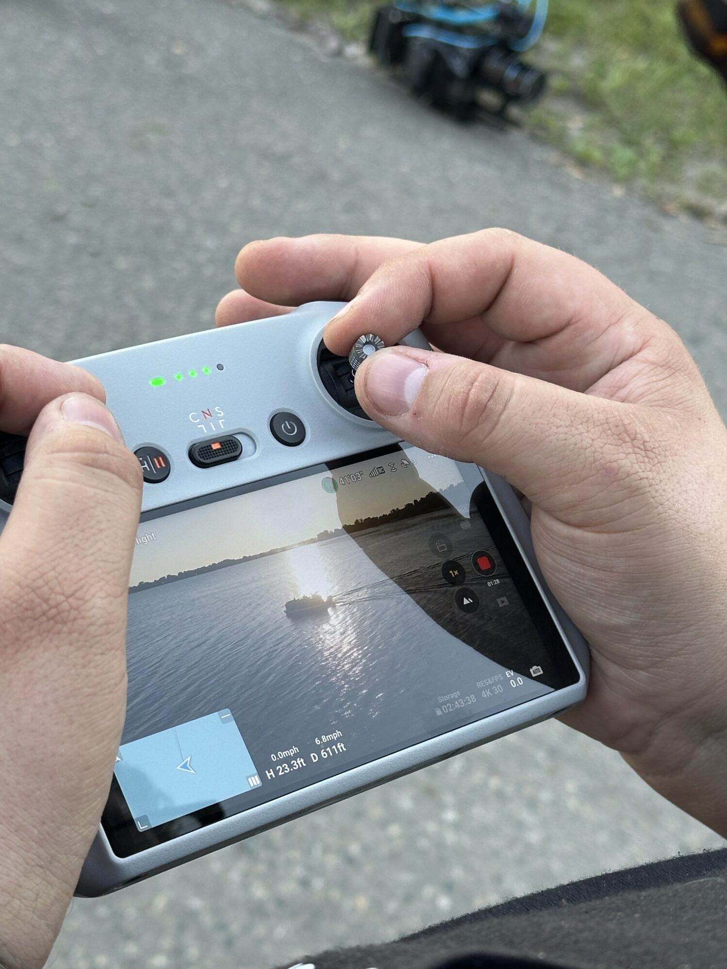 A person holding a drone remote. On the screen, you can see the drone footage that is being captured. On the screen, you see a boat in the middle of a lake during sunset.