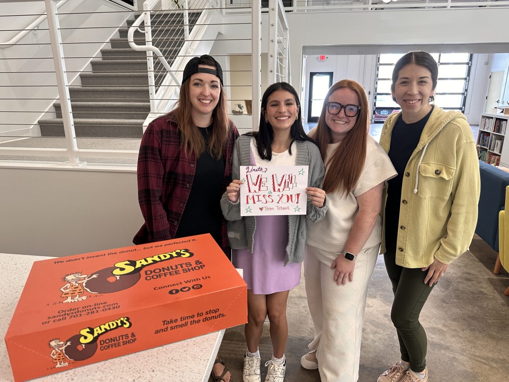A group of four women pose for a photo. One woman is holding a poster that reads “Lineth, we will miss you! Love Team Tellwell.” They are in a modern office setting. There is an orange box of Sandy’s Donuts in the foreground.
