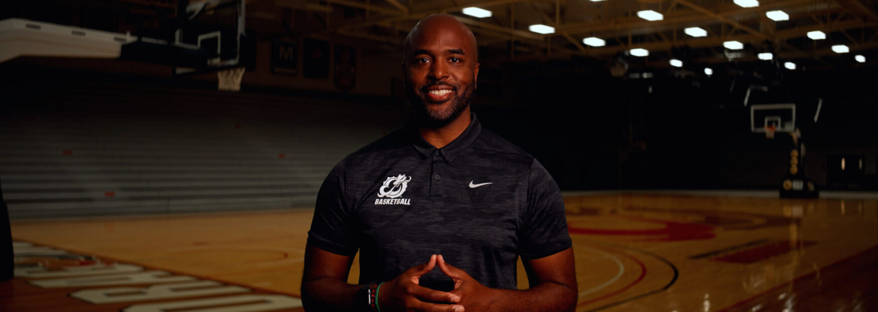 A man stands smiling on a basketball court, wearing a dark Nike polo shirt with a basketball team logo. The empty bleachers and hoop are visible in the dimly lit gym behind him.