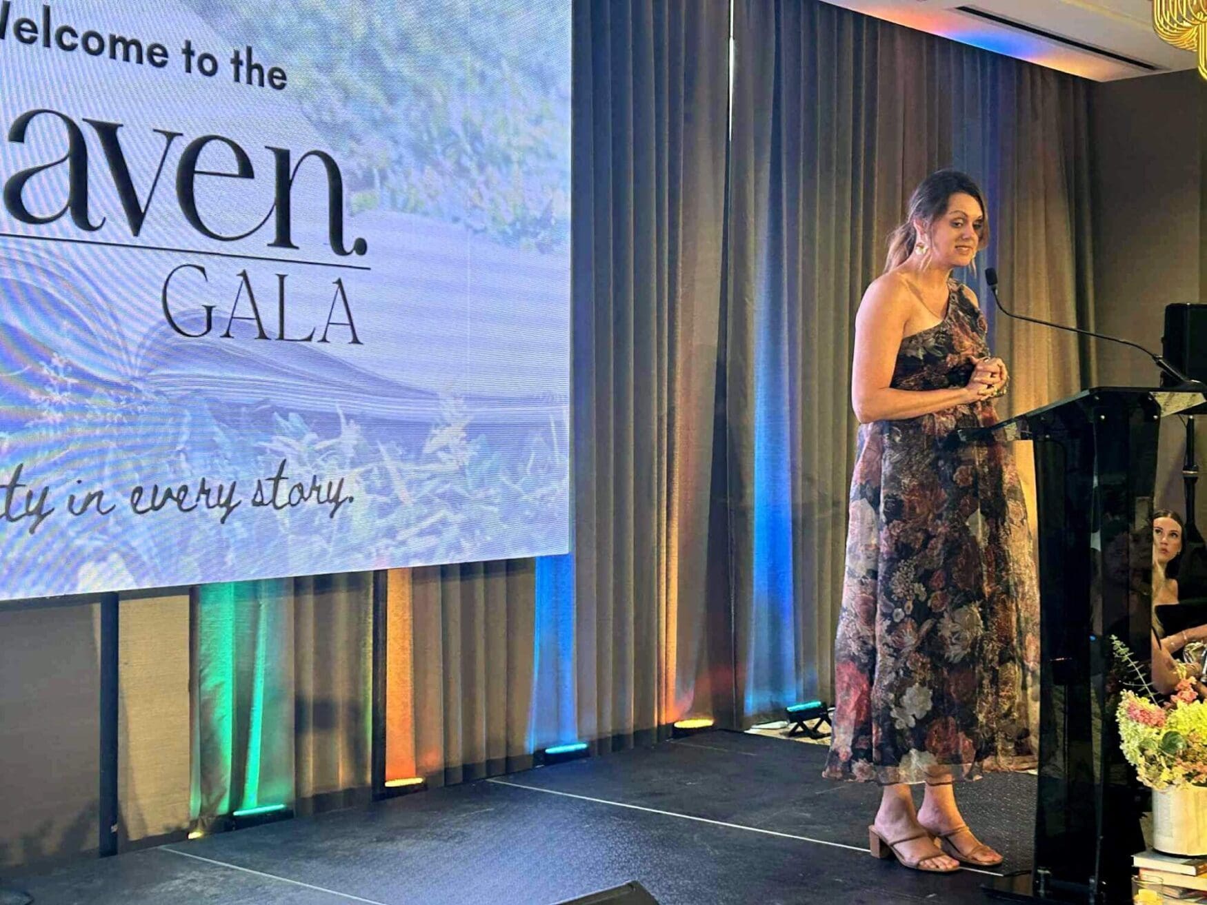 A woman in a formal dress stands at a podium on stage at an event. There is a screen that reads “Welcome to the Haven Gala.” The woman appears to be presenting to a crowd as she talks into a microphone.