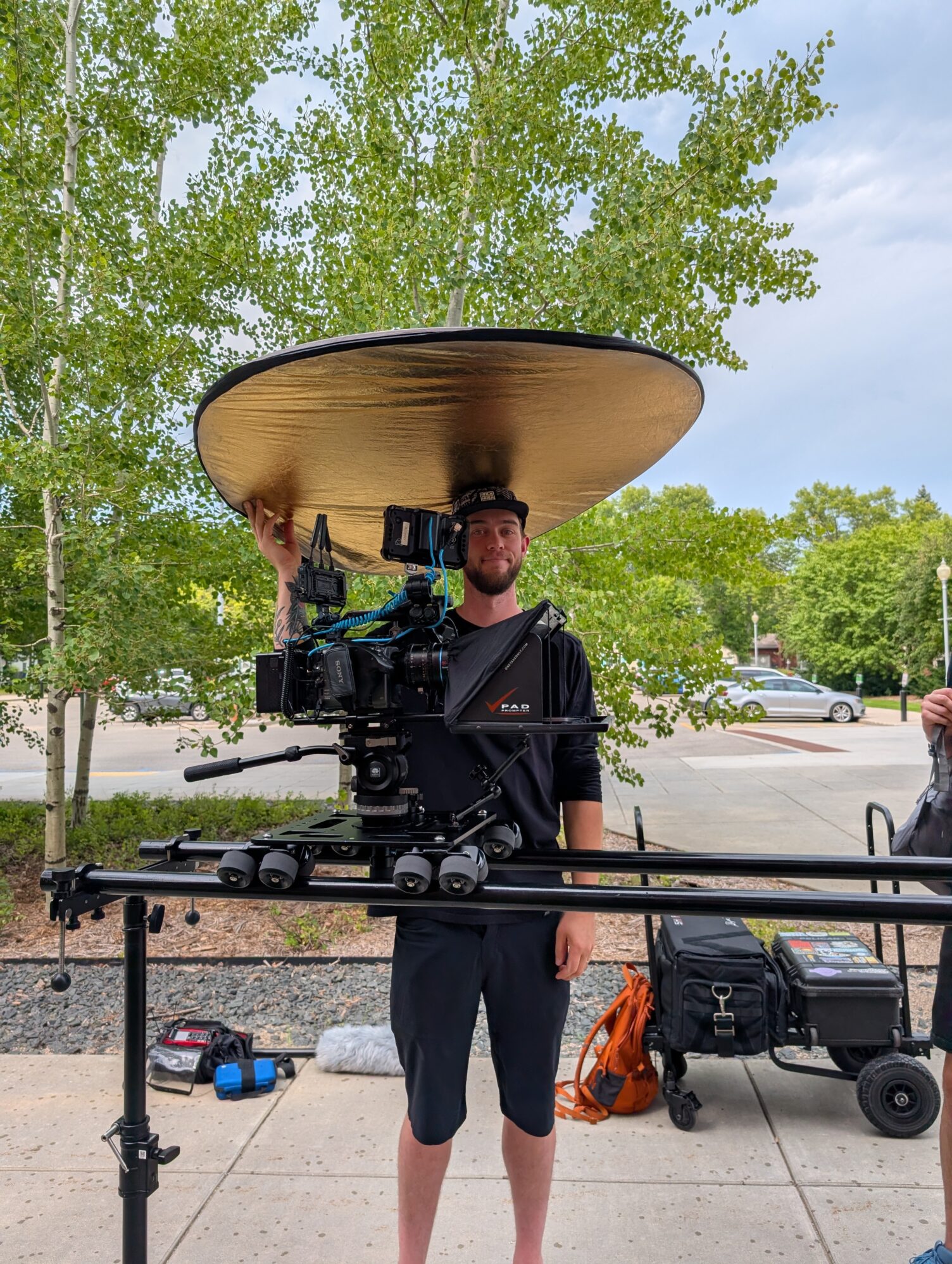 A man in a black shirt, shorts, and cap stands behind a professional camera mounted on a track slider. He is holding a large round gold reflector above his head, balancing it like a giant hat while smiling slightly. The setup is outdoors on a sidewalk, with leafy green trees and parked cars in the background. Equipment bags and cases are on the ground nearby.