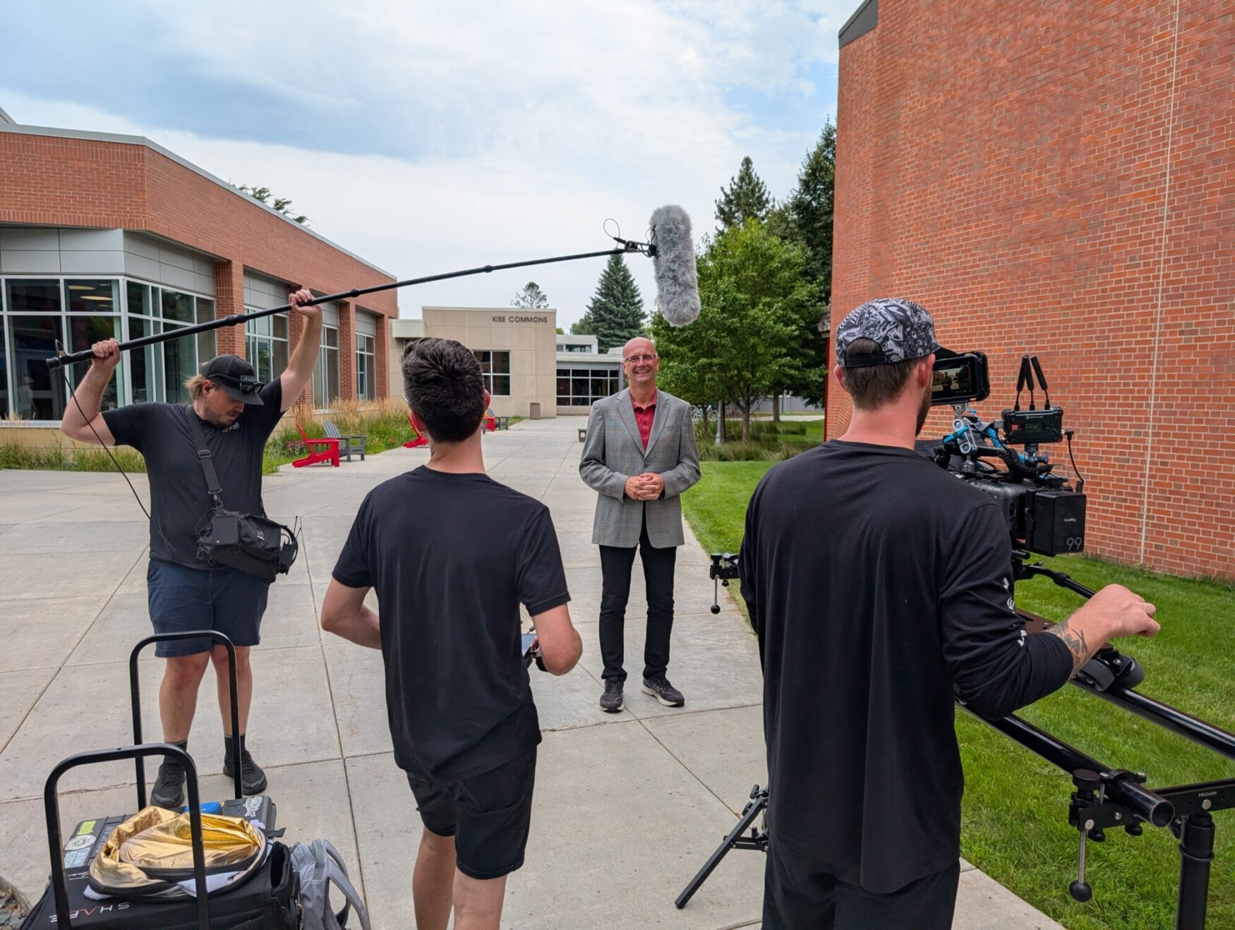 A small film crew is recording a man standing outside on a sidewalk between brick buildings. The man being filmed wears a gray blazer and glasses, smiling toward the camera. One crew member holds a boom microphone above him, another operates a large professional camera on a tripod slider, and a third stands nearby assisting. Equipment bags and gear are visible in the foreground, while the background shows a building labeled 'KISE COMMONS,' trees, and red outdoor chairs.