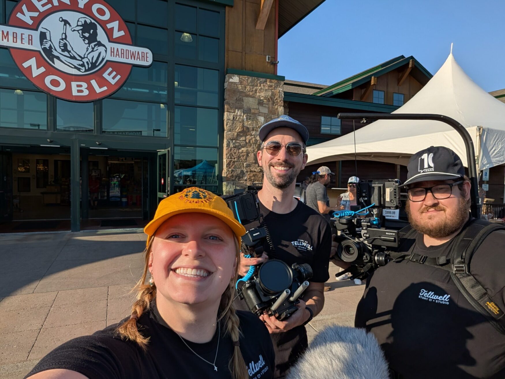 Two men and a woman all hold camera gear and smile for a photo. They are all wearing hats and black shirts that read "Tellwell Story Co." They are standing outside a large building that says "Kenyon Noble Lumber & Hardware."