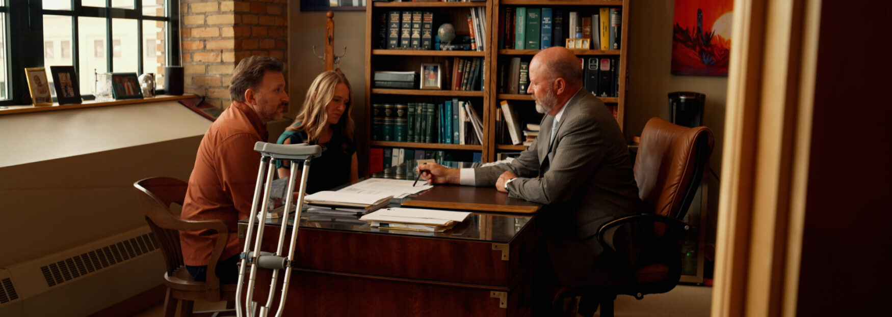 A lawyer in a gray suit sits at a wooden desk speaking with a man and woman seated across from him. The office is filled with bookshelves and legal books, with framed photos on the windowsill. A pair of crutches leans against the desk, suggesting a personal injury case.