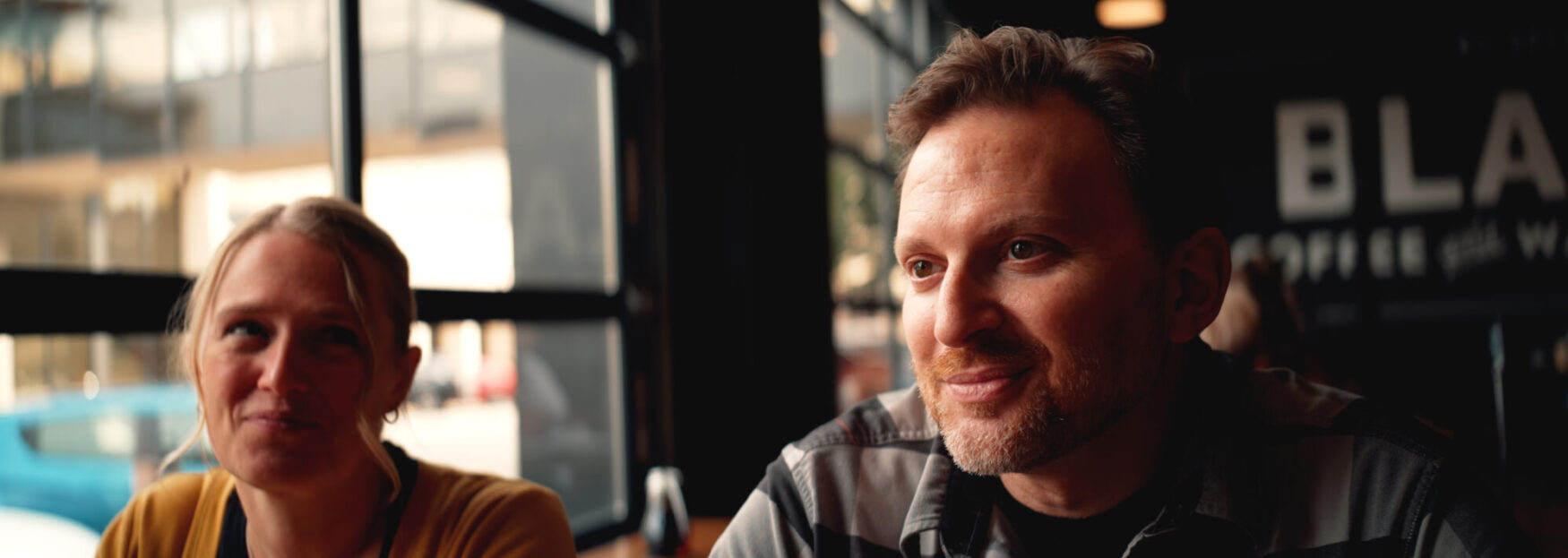 A man with light facial hair smiles while sitting in a coffee shop next to a woman who is softly smiling. The window behind them lets in natural light, with blurred city reflections visible.
