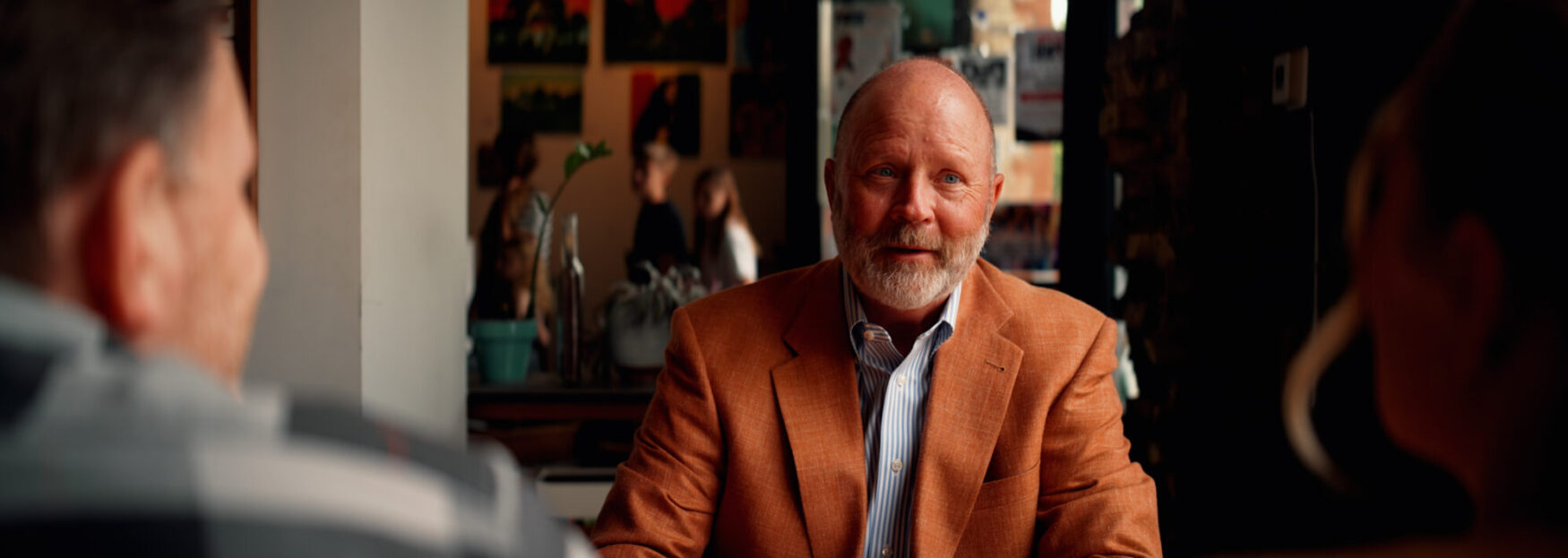 A bearded man in an orange jacket sits at a table in a coffee shop, speaking with two people whose faces are blurred in the foreground. Behind him are colorful paintings and people moving in the background.