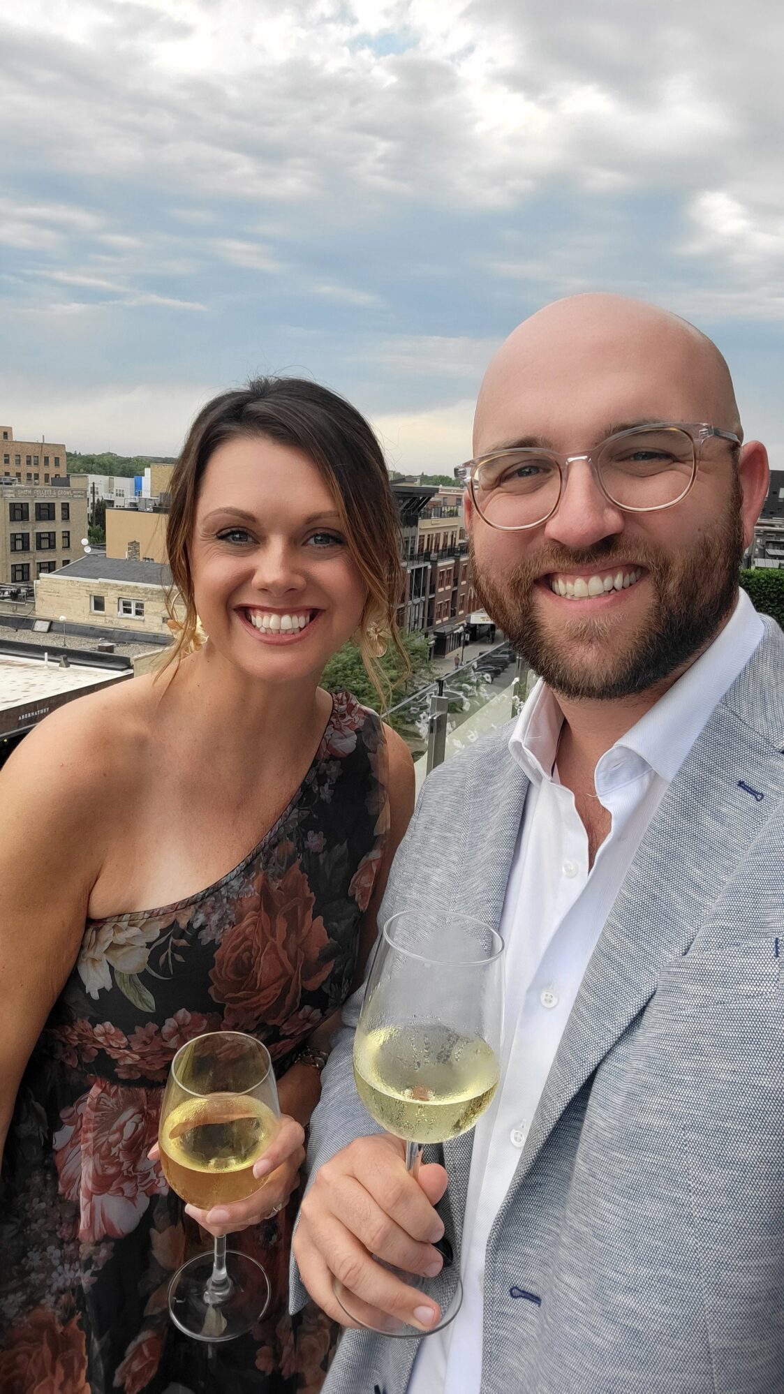 A man and a woman smile for a photo. They are both dressed formally and holding glasses of white wine. They appear to be on the terrace of a tall building, with the city skyline in the background.