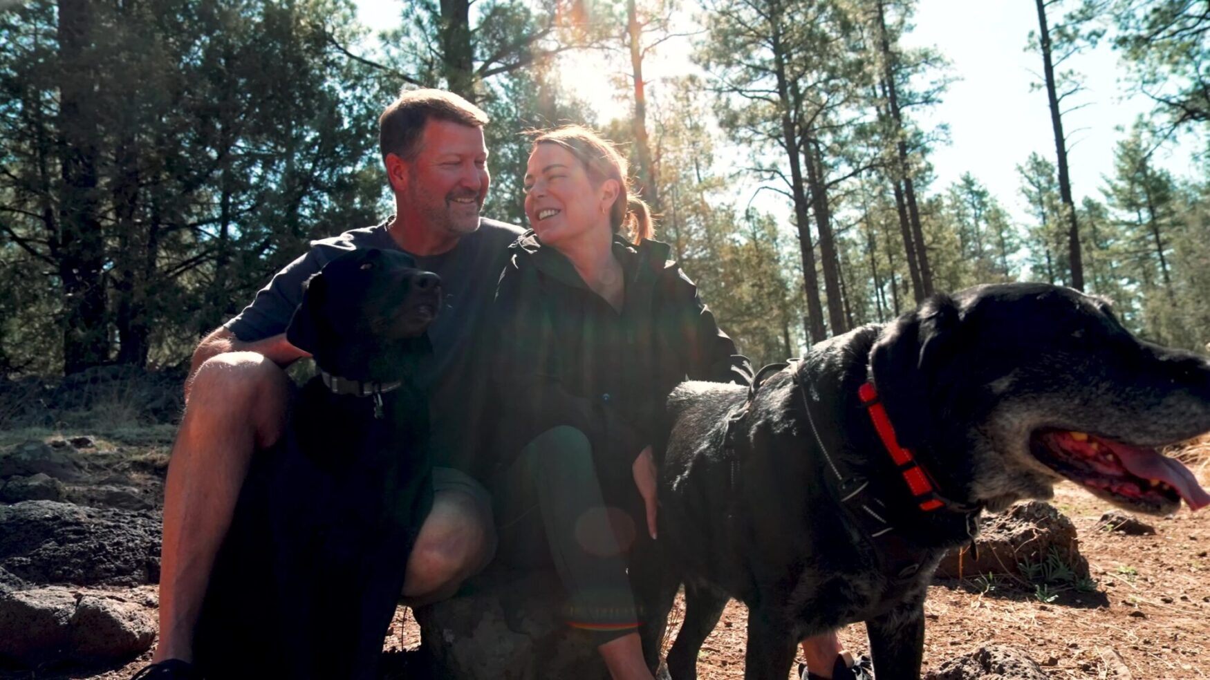 A man and a woman sit in the woods with their elderly black lab. The dog is wearing a red collar and the sun is shining behind them.