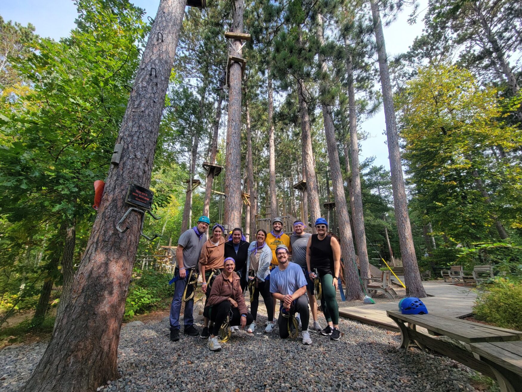 A group of people wearing helmets and harnesses gathered at the base of tall trees in a ropes course area, ready for adventure.