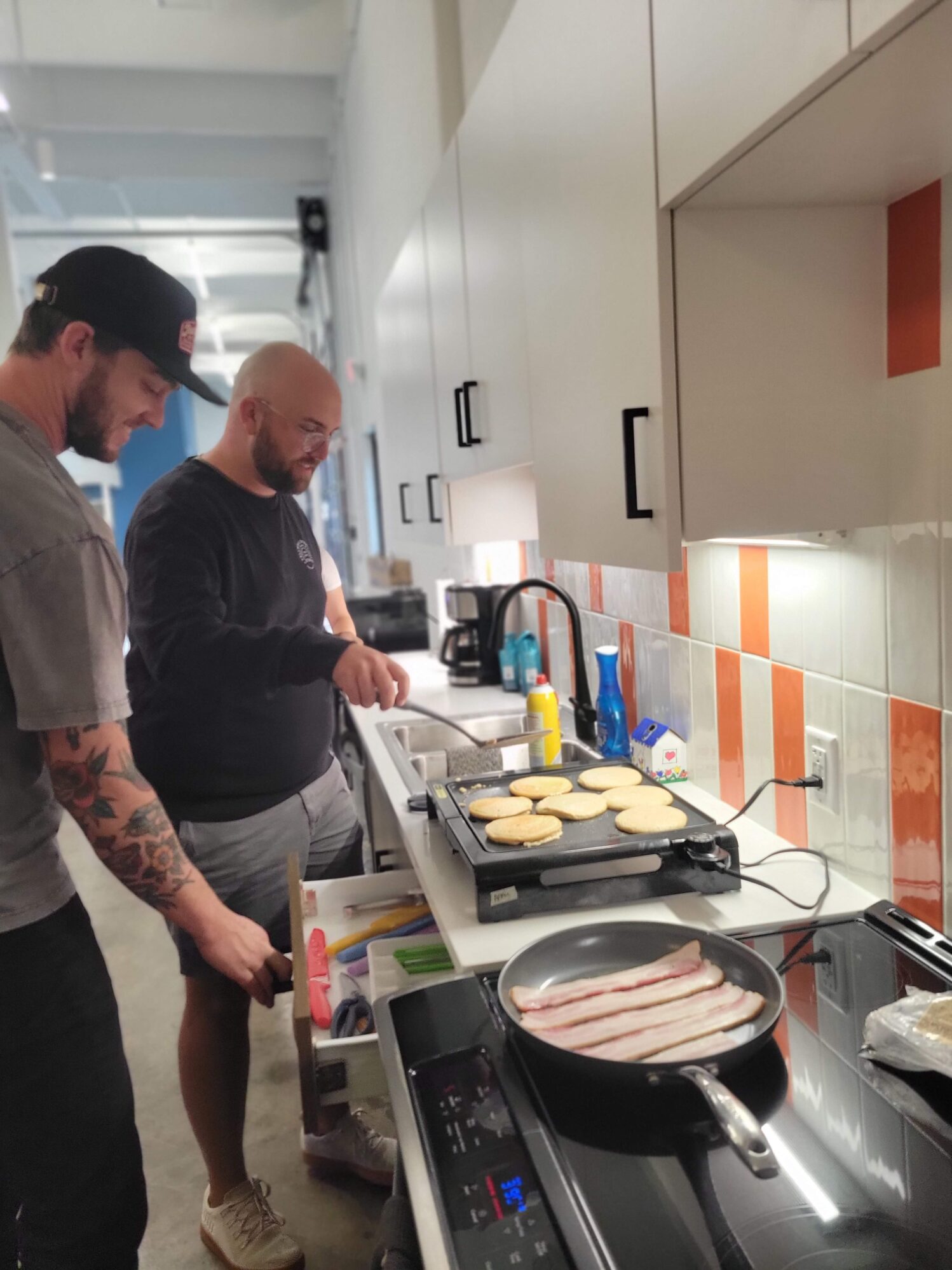 Two team members cooking pancakes on a griddle and bacon in a pan in the Tellwell kitchen.