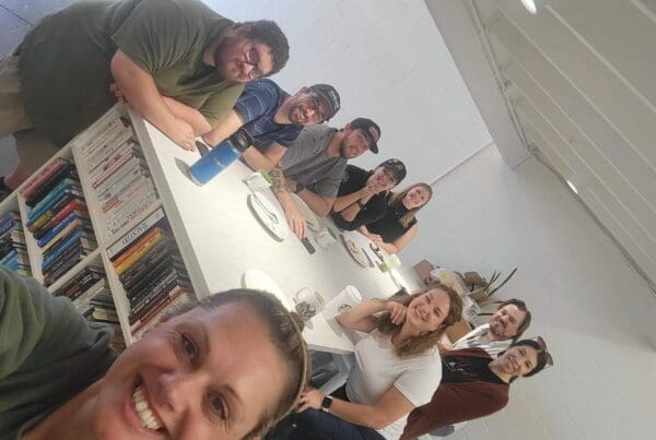 A group of smiling Tellwell team members sitting together around a long white table, mugs and plates in front of them.