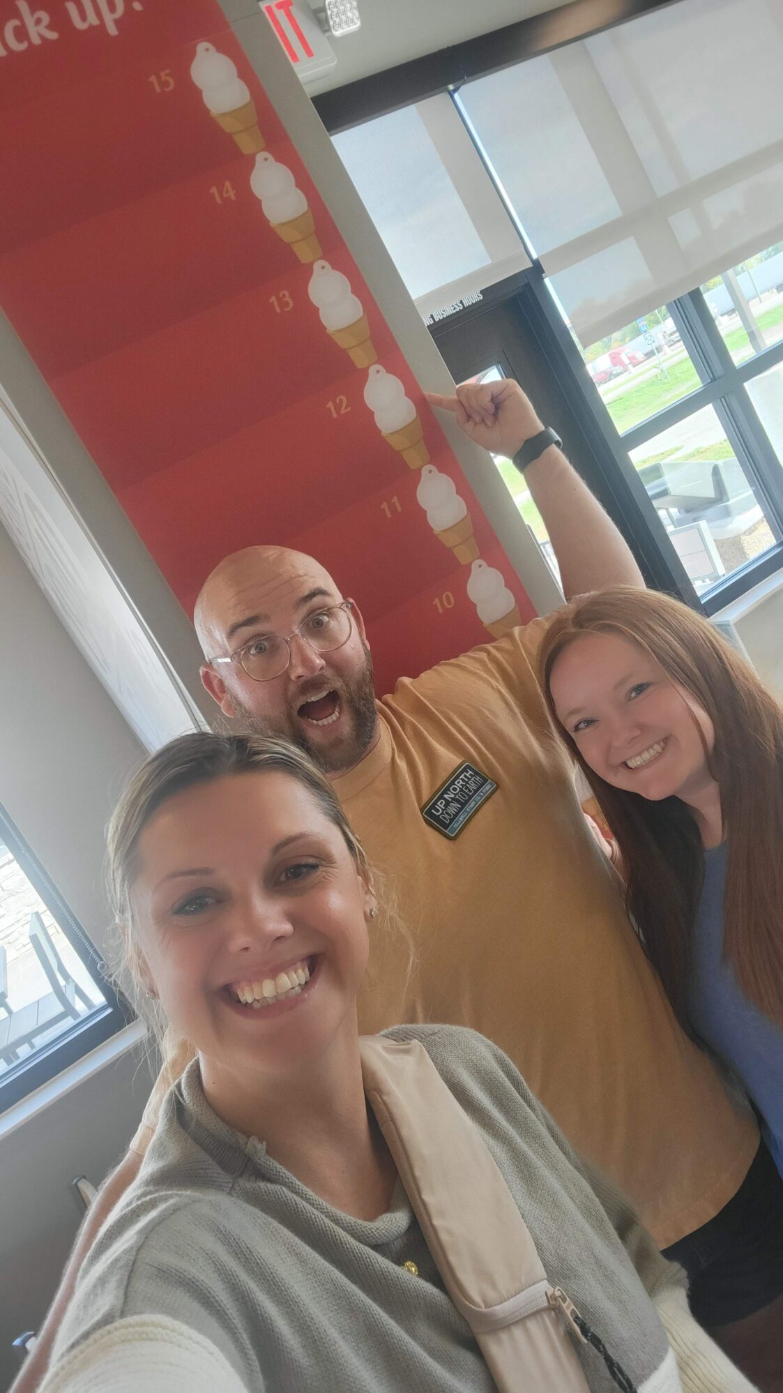 Three smiling people taking a selfie indoors. Behind them is a large red chart on the wall showing ice cream cones stacked with numbers from 10 to 15. The man in the middle, wearing glasses and a tan shirt, is pointing excitedly at the cone labeled 12. The woman in front is holding the camera and smiling widely, and another woman with long red hair is smiling next to the man.
