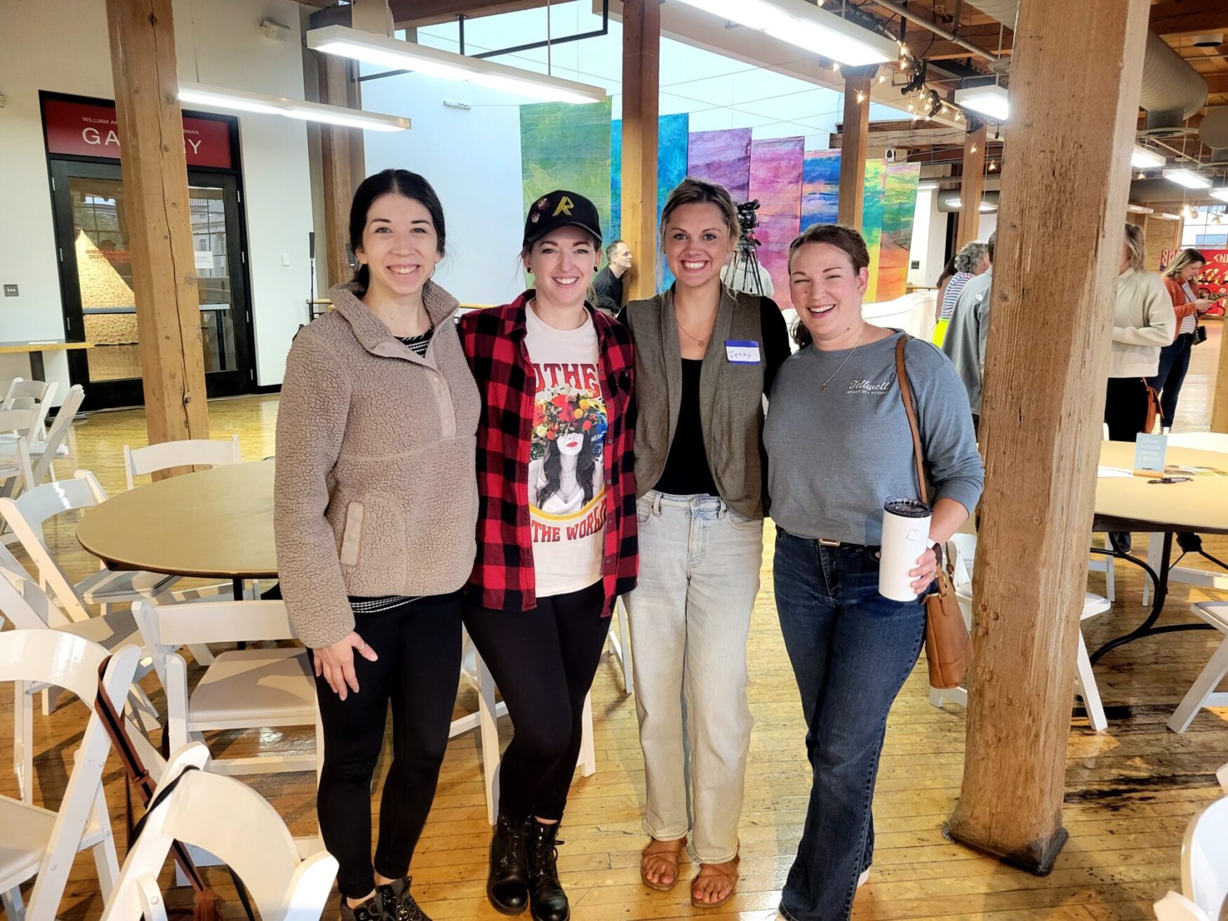 Four women stand close together and smile for a photo inside a bright, open event space with wooden beams and colorful artwork on the wall. They are dressed casually, with round tables and white chairs set up around the room, and other people visible in the background.
