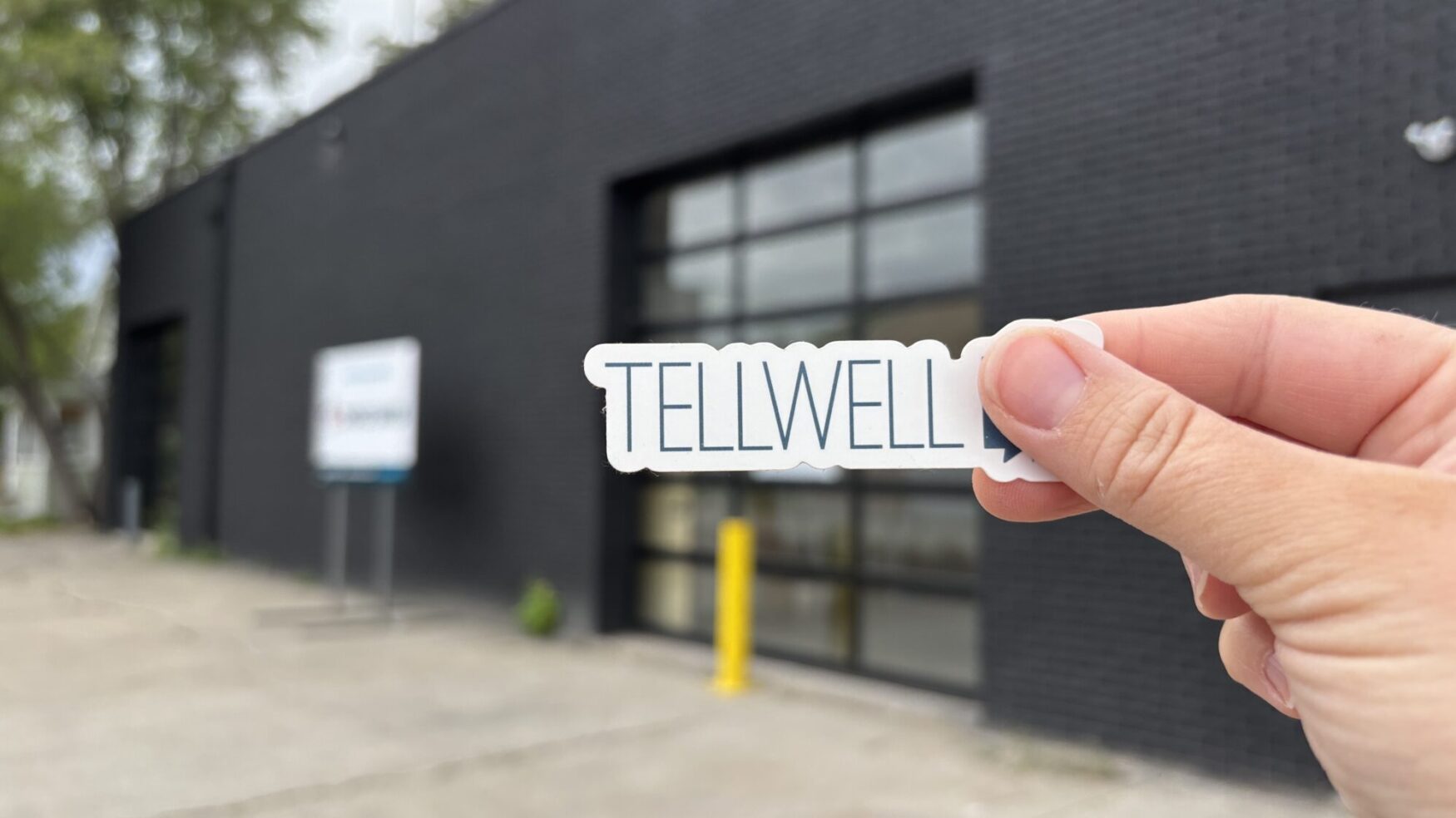 A hand holding a Tellwell sticker in front of the new black-brick Tellwell office building.