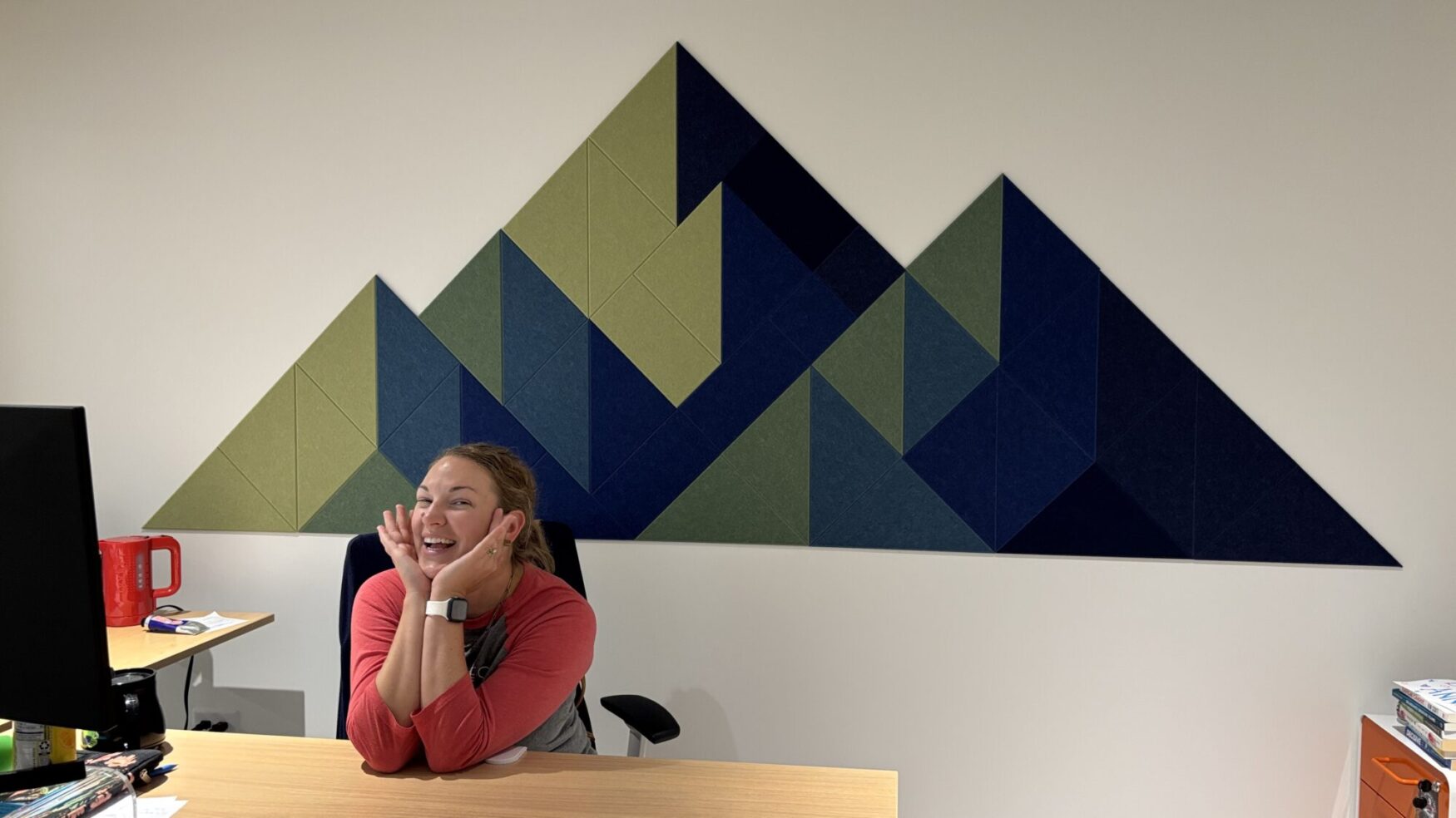 A woman sitting at a desk smiles playfully with her hands resting under her chin. She wears a red and gray shirt and a smartwatch. Behind her on the wall is a large geometric mural made of triangular panels in shades of green and blue, arranged in the shape of mountains. The desk has papers, a computer monitor, and a red mug, with books and other items on a side cabinet.