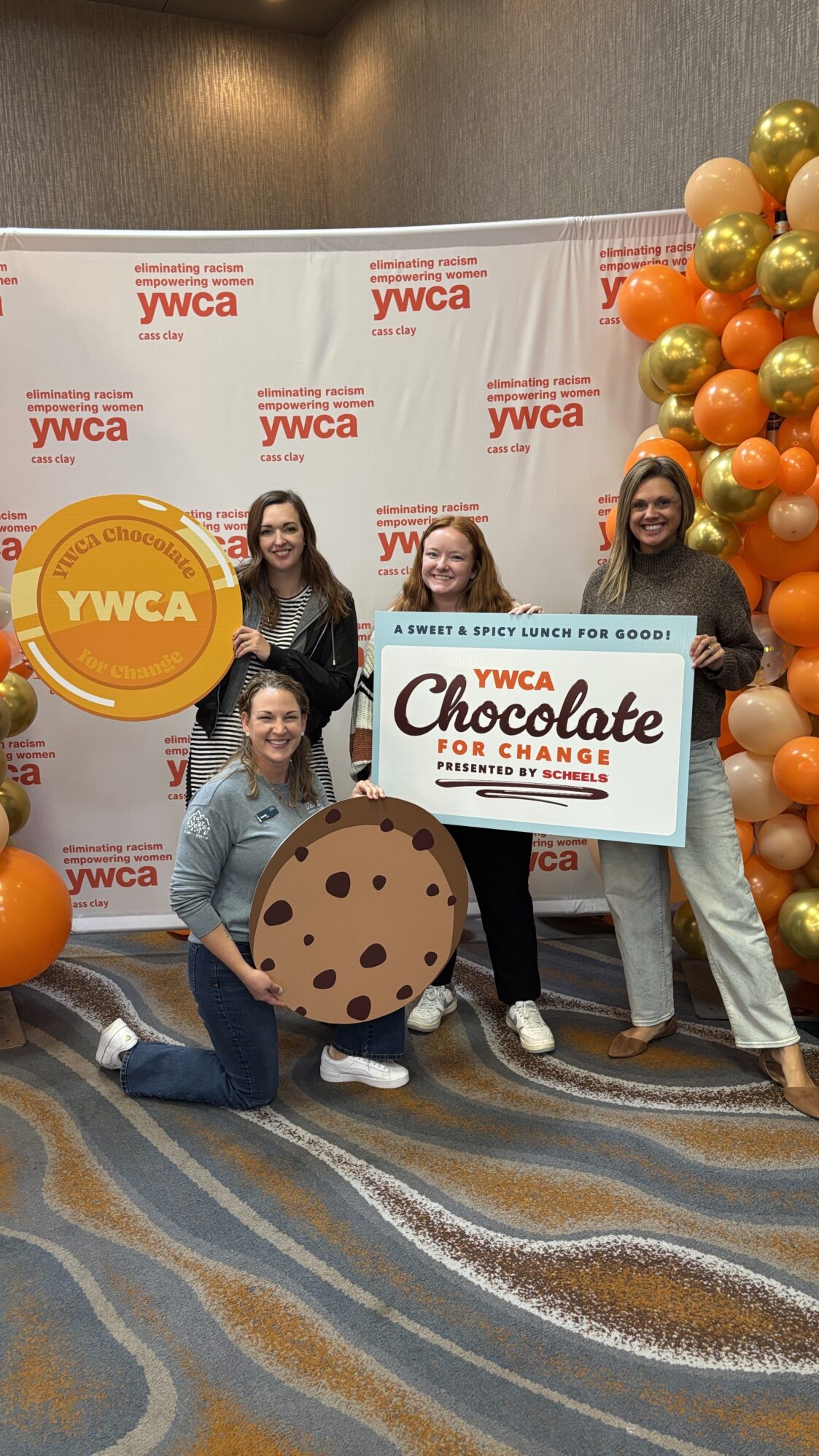Four women pose together in front of a YWCA Cass Clay step-and-repeat backdrop with the slogan “eliminating racism, empowering women.” They are smiling and holding oversized props, including a large cookie, a circular “YWCA Chocolate for Change” token, and a rectangular sign that reads “YWCA Chocolate for Change: A Sweet & Spicy Lunch for Good! Presented by Scheels.” To the right, there is a festive balloon display in orange, gold, and cream colors.