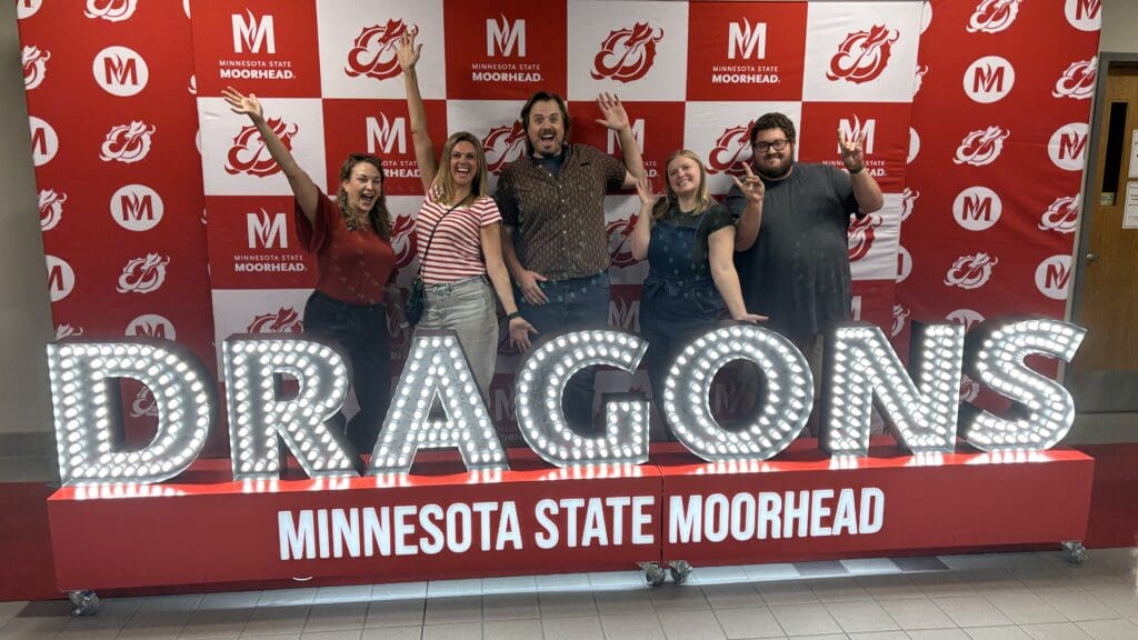 Five people pose enthusiastically in front of a red-and-white Minnesota State University Moorhead step-and-repeat backdrop. They stand behind large illuminated letters spelling “DRAGONS,” with “MINNESOTA STATE MOORHEAD” displayed below.