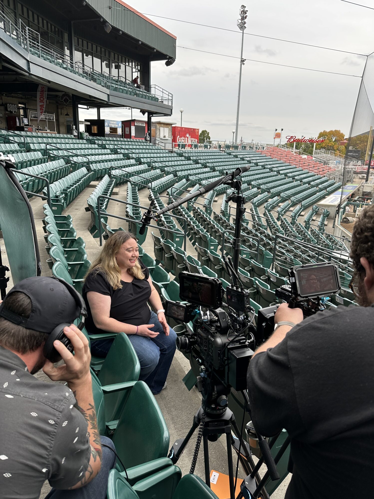 A woman sits in the stands of a mostly empty stadium, smiling as she is filmed for an interview. Professional cameras and microphones are set up in front of her, with crew members operating the equipment. Rows of green stadium seats and part of the concourse are visible in the background.